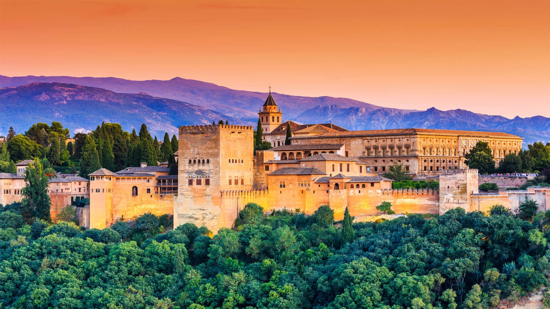 View of the Alhambra in Granada, Spain, a fortified palace surrounded by forest with mountains in the background.