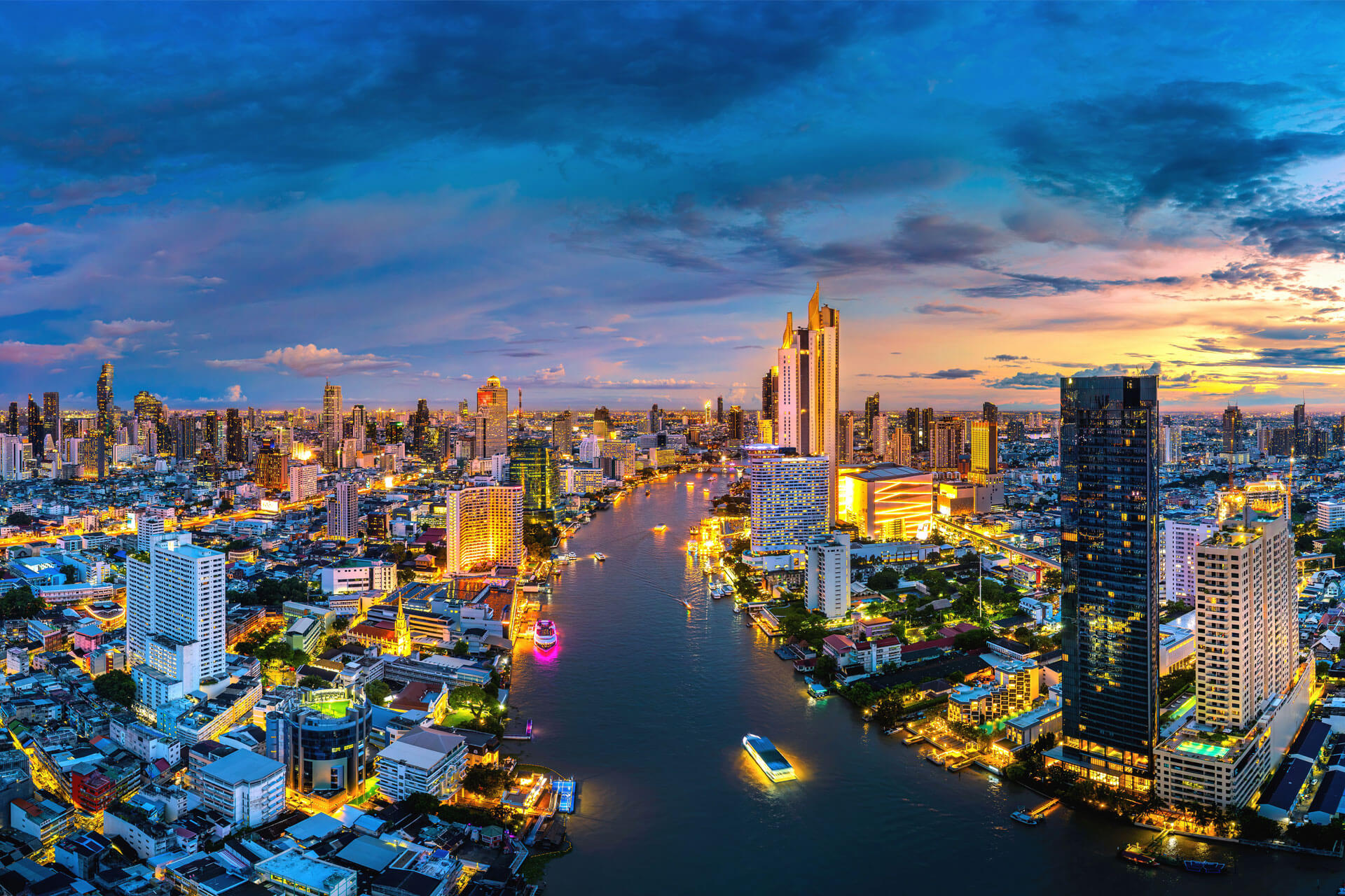 Aerial view of Bangkok at dusk, with illuminated skyscrapers and the Chao Phraya River winding through the city.