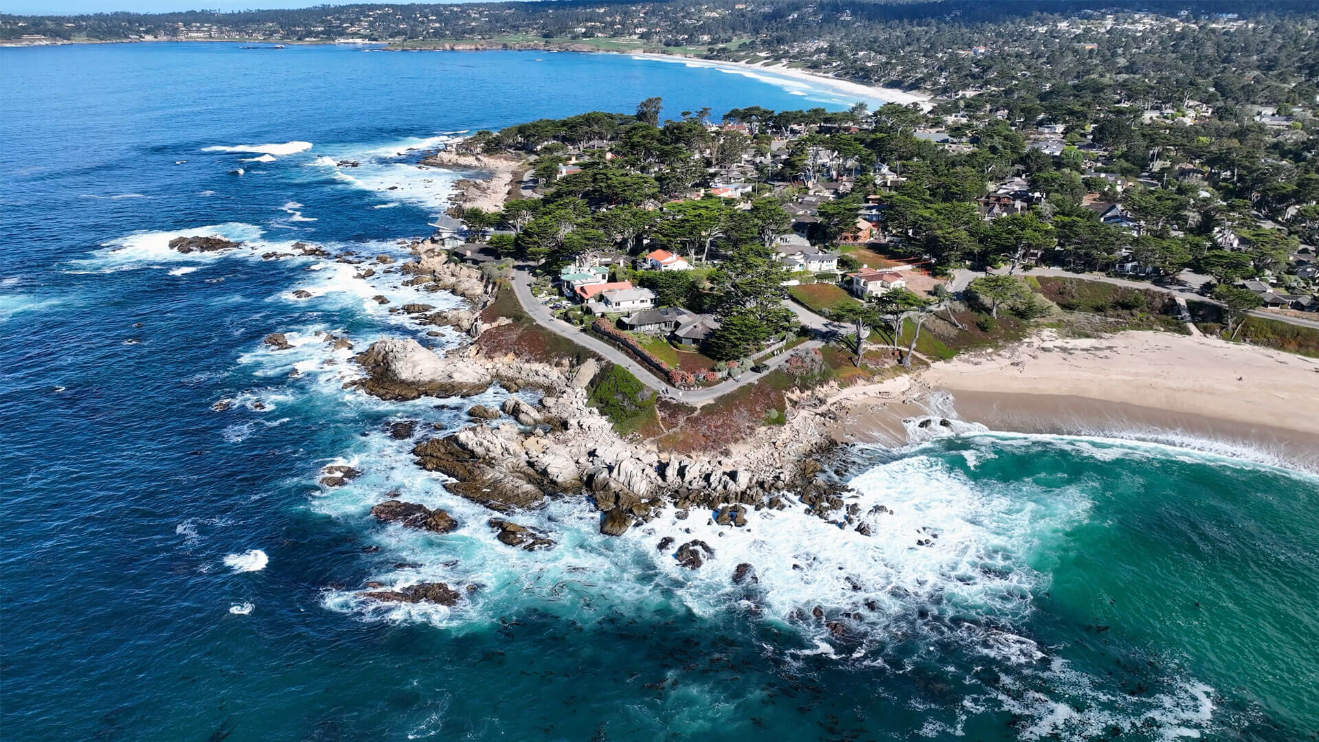 Aerial view of Carmel-by-the-Sea with rocky cliffs, sandy beaches, turquoise waters, and towering cypress trees.