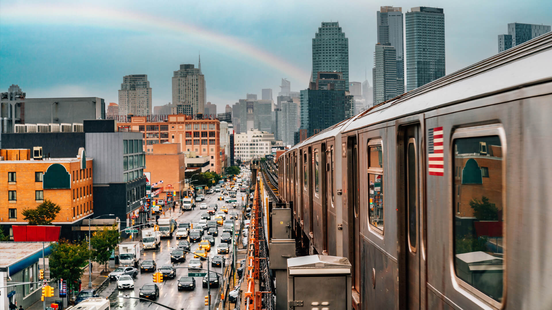 Queens elevated subway in New York, overlooking a street with cars and yellow taxis under a cloudy sky and a rainbow.