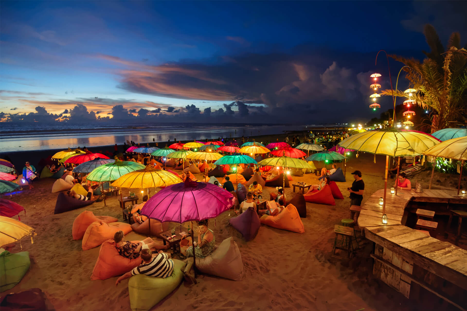 Colourful illuminated umbrellas on Bali’s lively Seminyak beach at night, with people relaxing on bean bags.