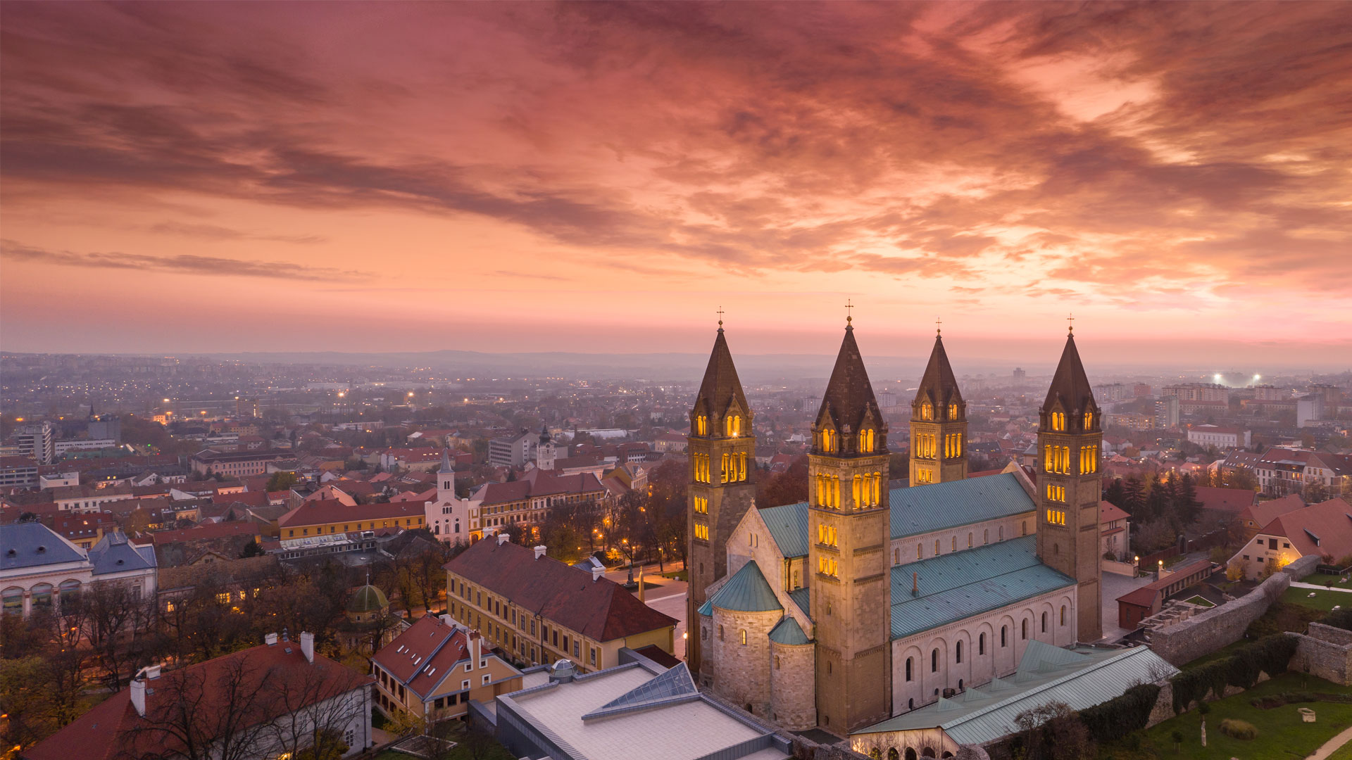 Vue aérienne de Pécs, Hongrie, le soir, avec la cathédrale Saint-Pierre-et-Saint-Paul illuminée et un ciel rosé se reflétant sur la ville.