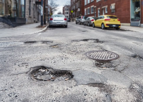 Gros plan sur un nid-de-poule rempli d’eau dans un quartier urbain. Des voitures stationnées sont visibles des deux côtés de la rue.