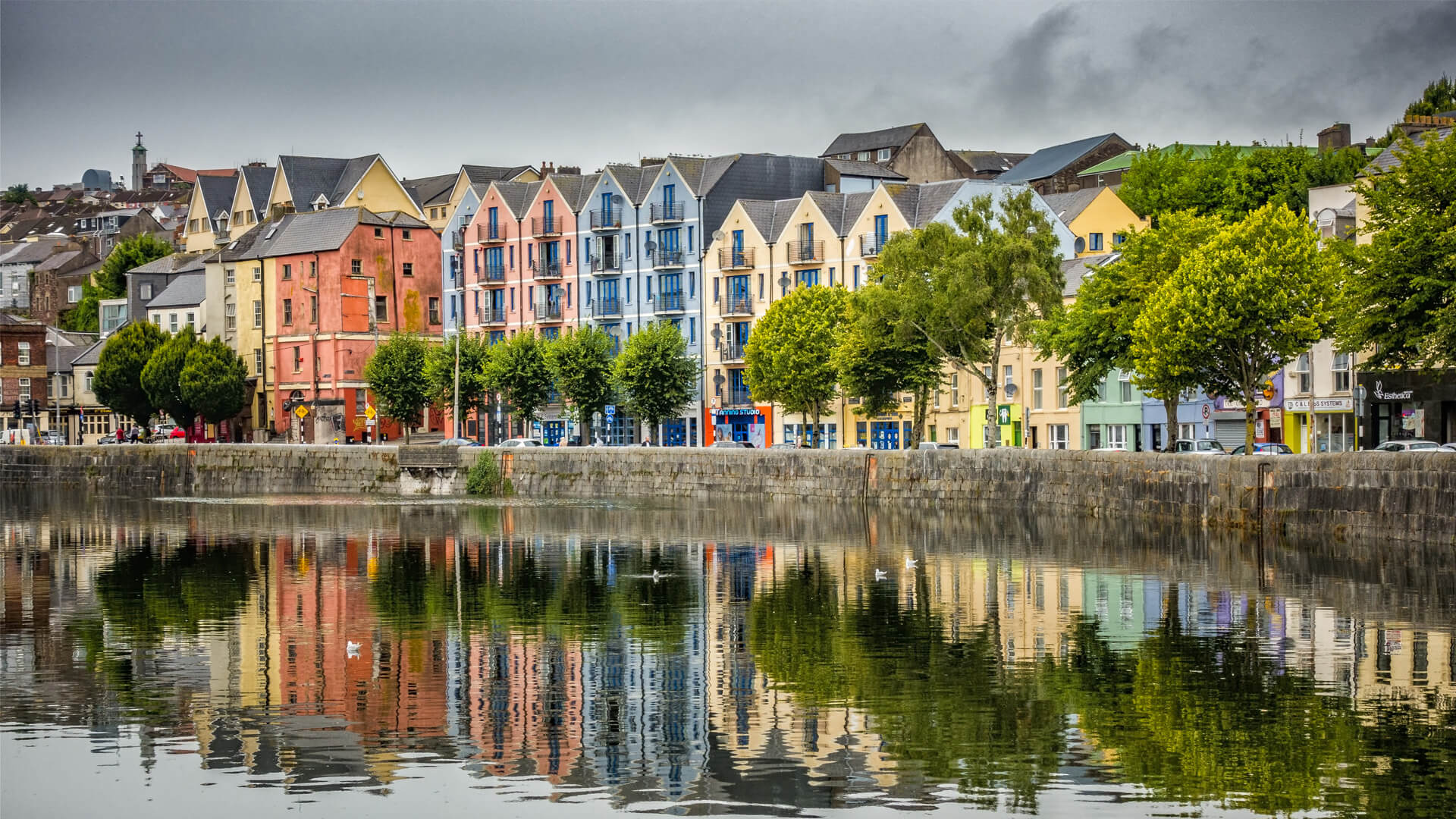 Colourful houses in Cork along the river, lined behind lush green trees, reflected in the water under a grey sky.