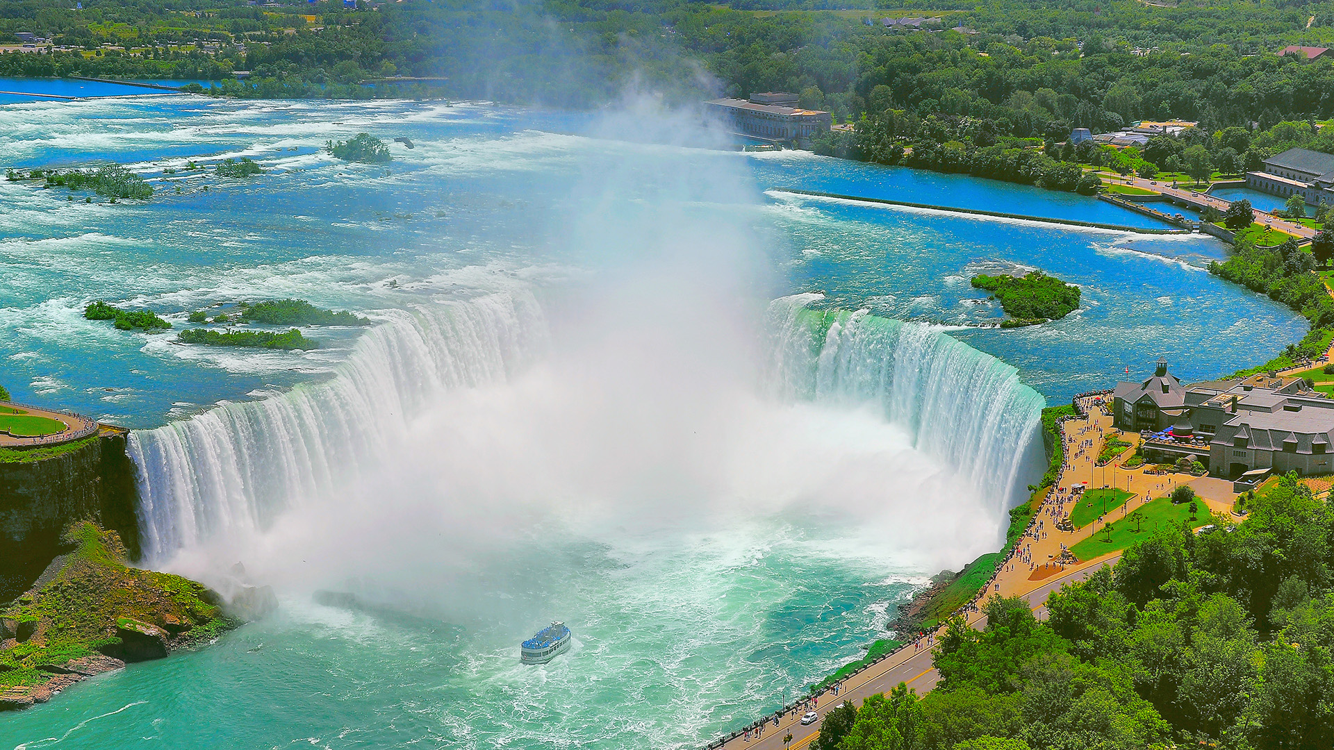 Aerial view of Niagara Falls, Ontario, with a sightseeing boat, mist rising from the falls, and lush greenery under a blue sky.