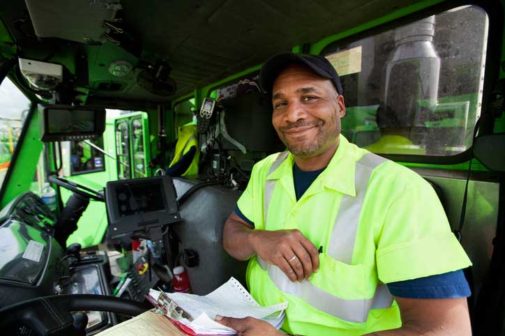 Driver dressed in high-vis clothing sitting in sanitation vehicle smiling at the camera.