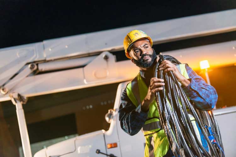 Main in hardhat and vest caring a large coil of cord over his shoulder