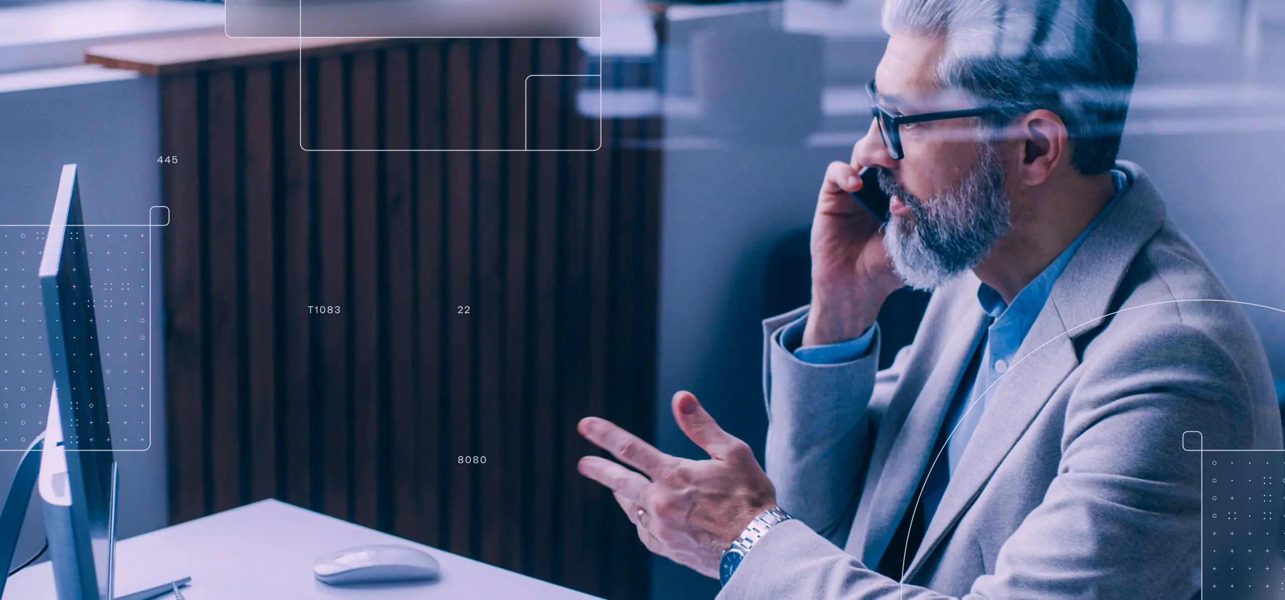 Gray-haired man in light blazer holds smartphone to ear and gestures at desk beside a computer, with faint UI overlay lines