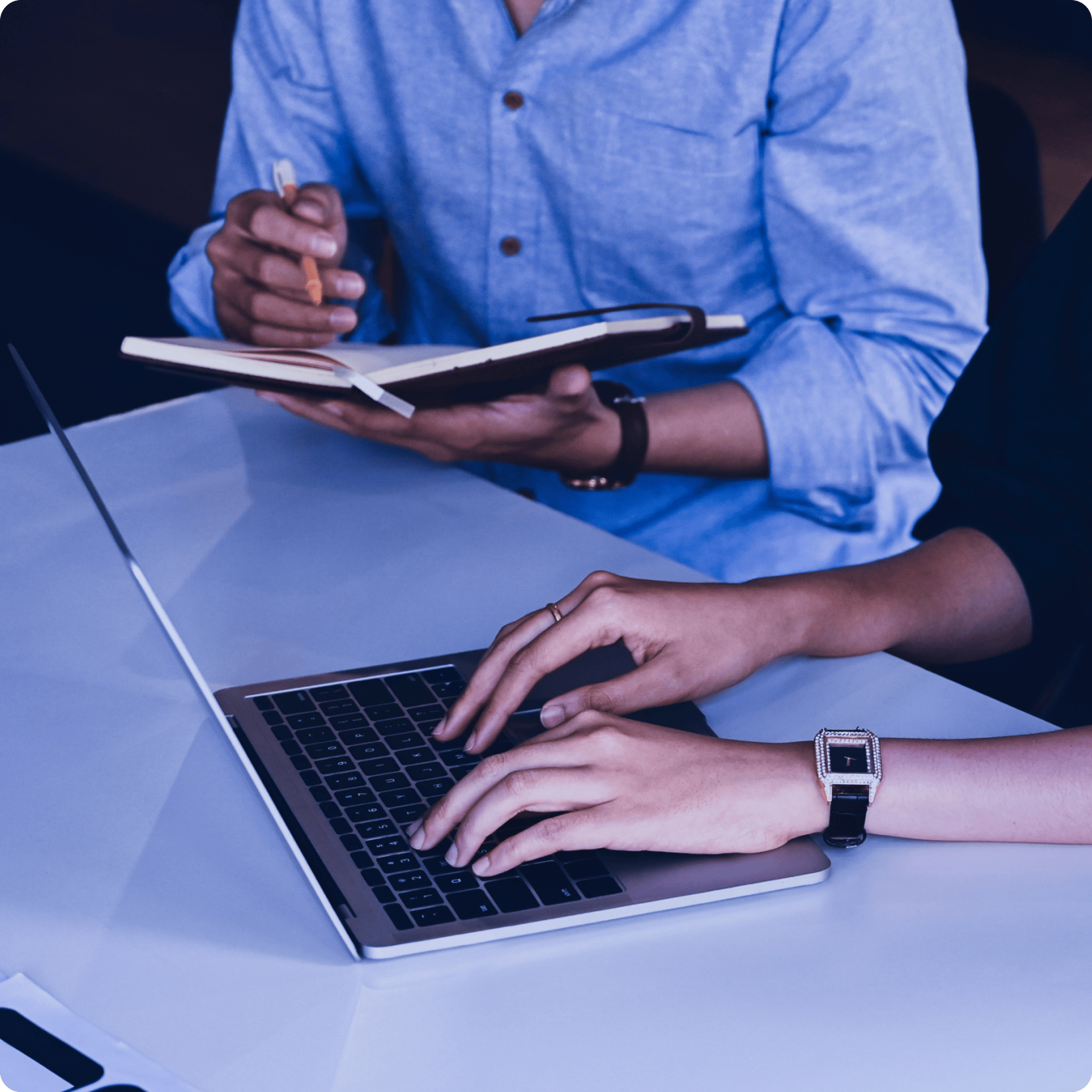 Two people seated at a table, one writing in a notebook and one typing on a laptop