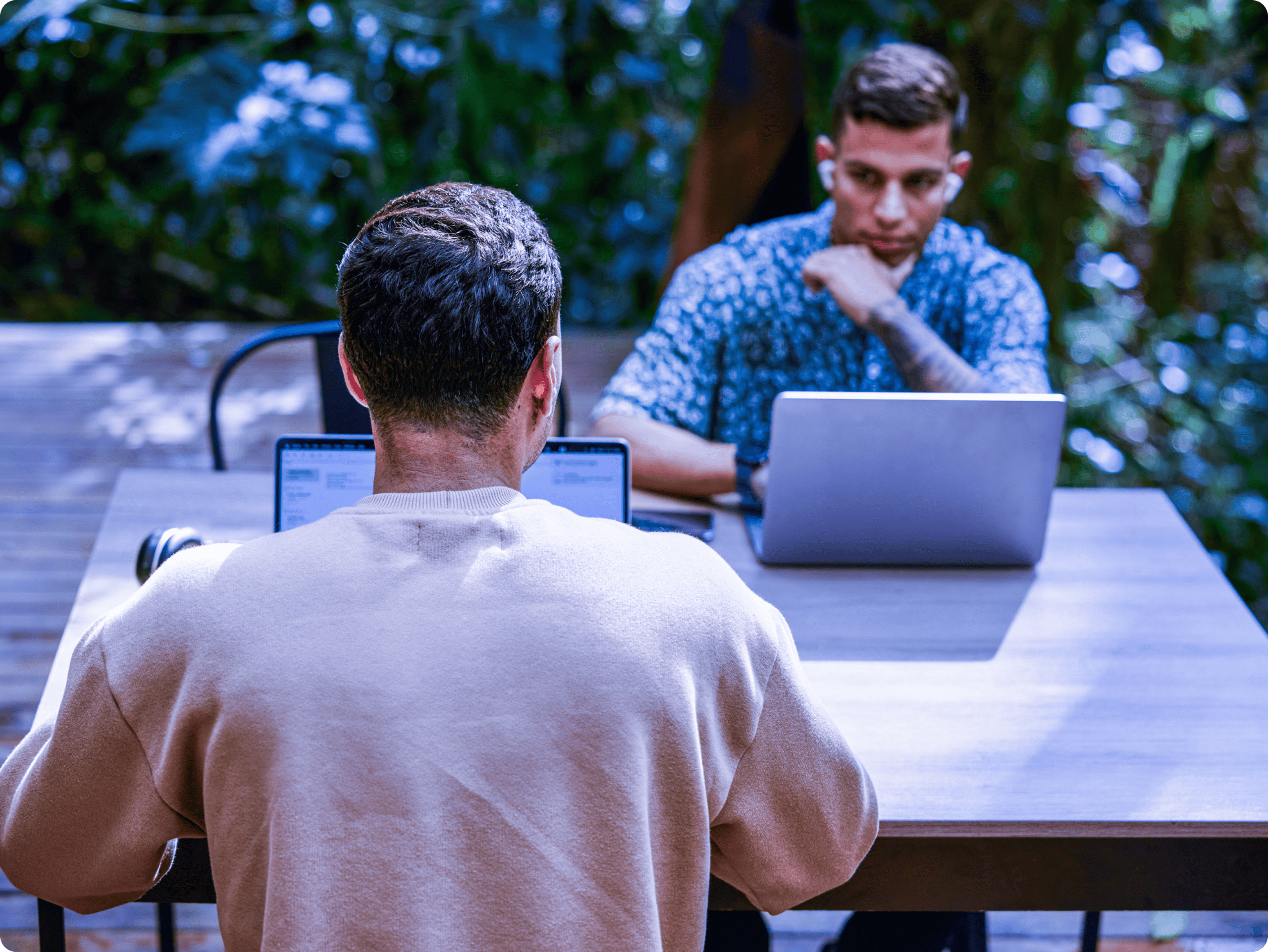 Two men working on open laptops at an outdoor patio table with headphones nearby, surrounded by green foliage