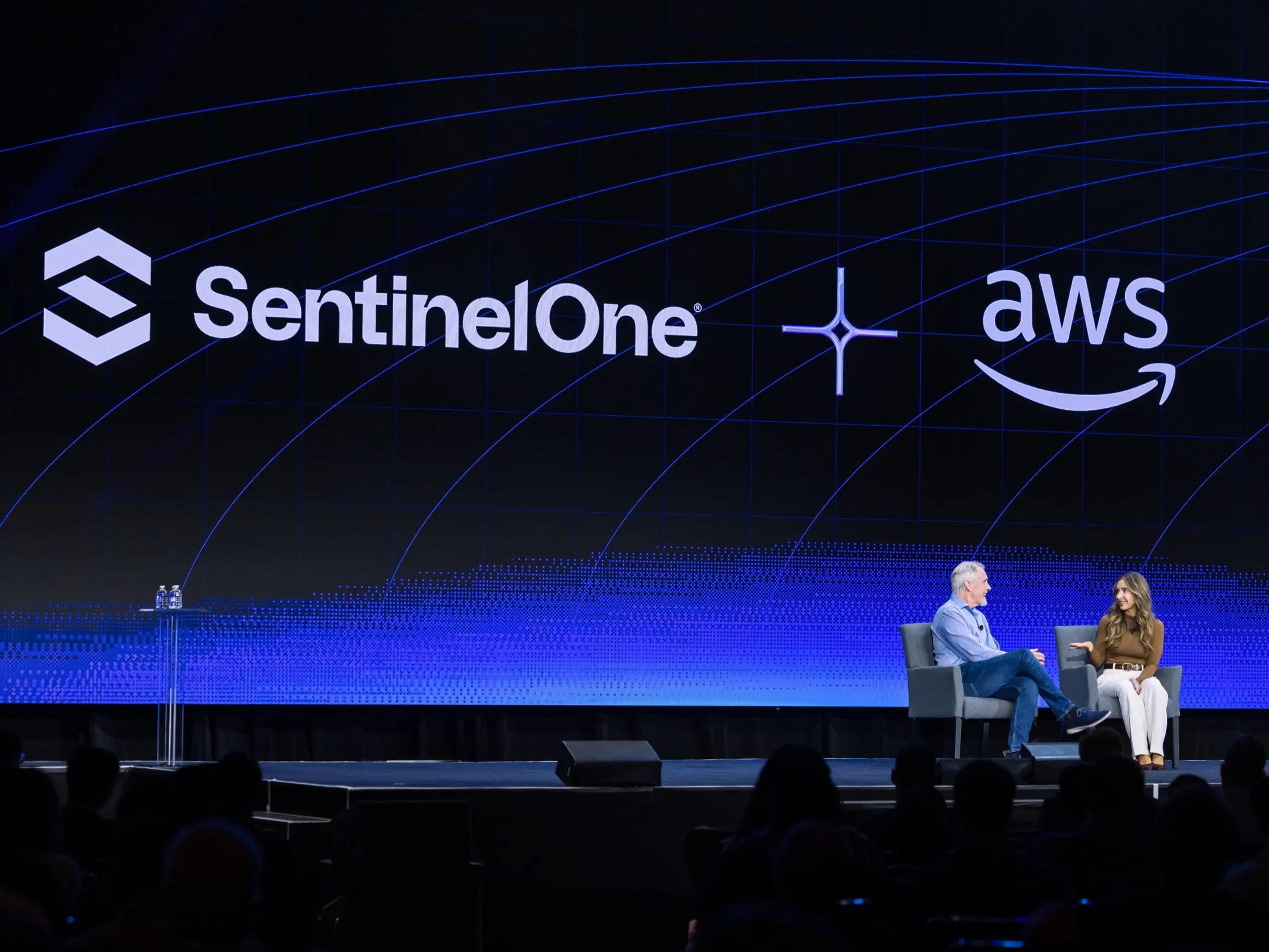 Stage with dark LED screen showing SentinelOne and AWS logos, blue arcs and band, plus symbol; two people in chairs and clear podium