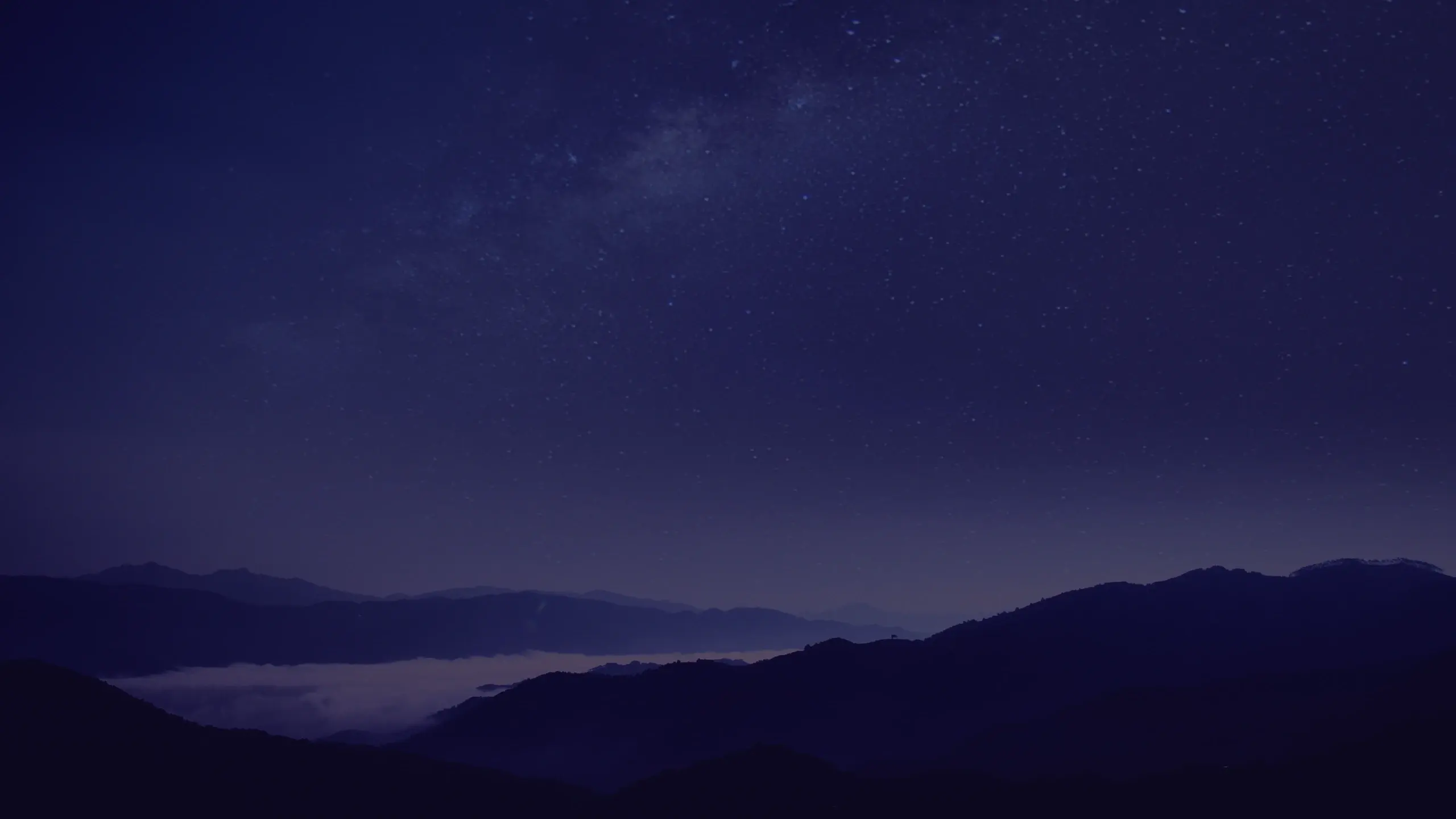 Night landscape with deep blue and purple starry sky, layered dark mountain silhouettes, and a hazy valley band