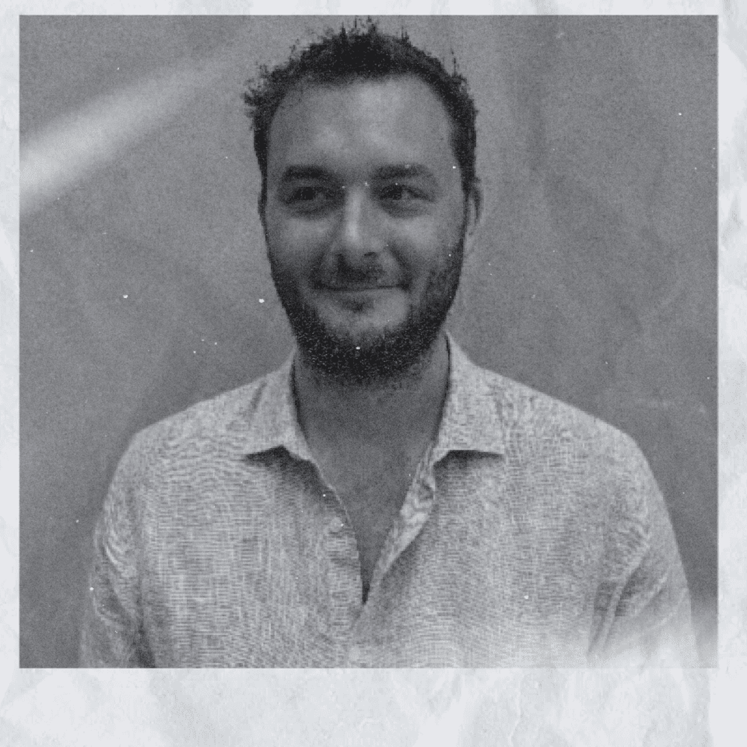 Grayscale chest-up portrait of a bearded man in a light open-collar shirt, faint smile, plain blurred background