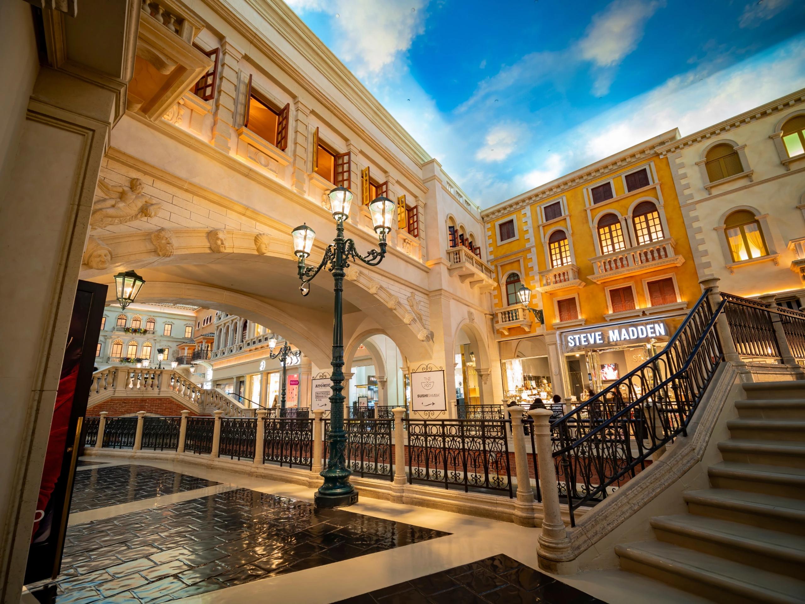 Covered European shopping walkway with painted blue sky, lamppost, staircase, and storefront sign reading STEVE MADDEN