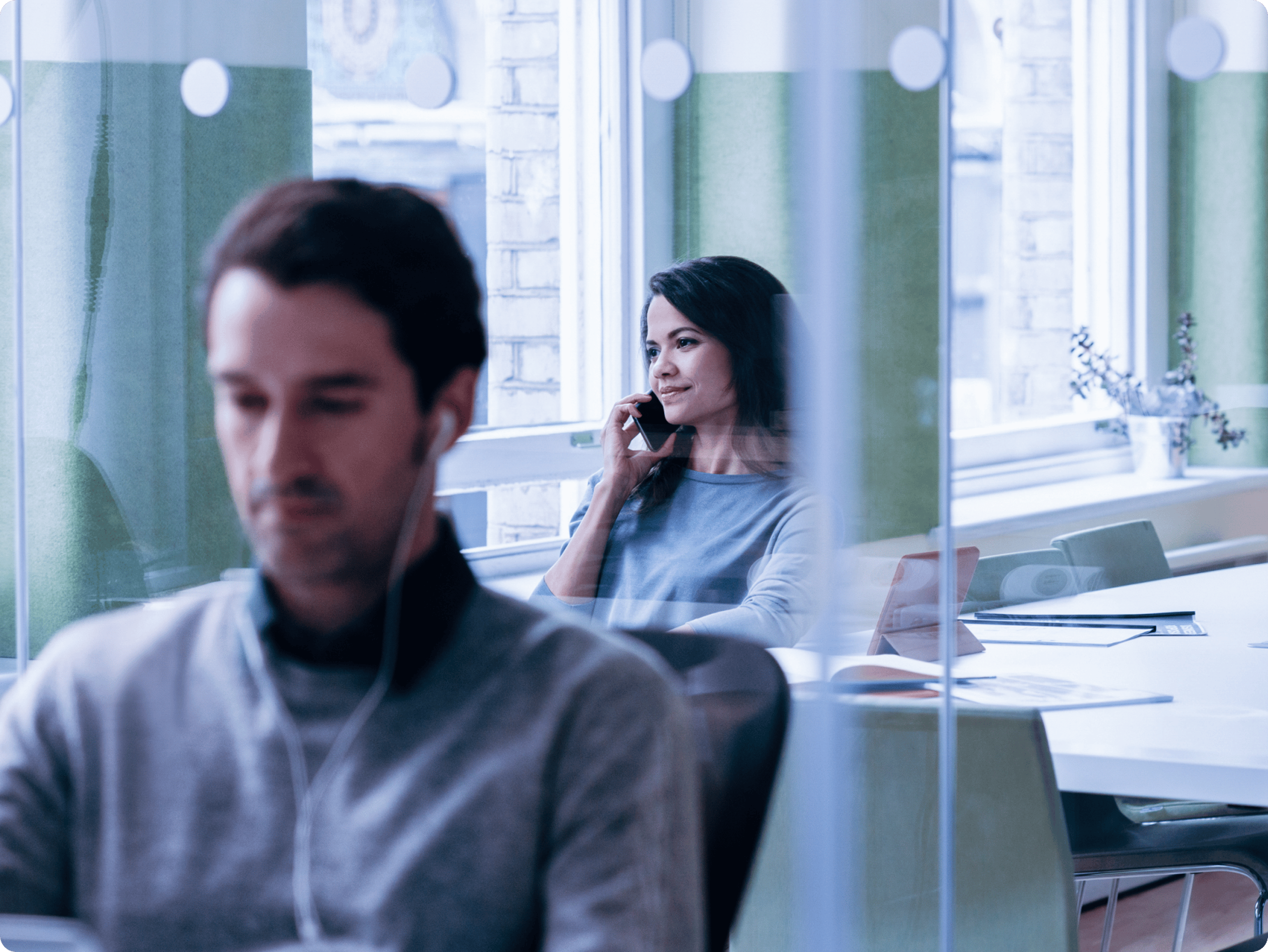 Office interior seen through a glass partition; two people seated at desks, daylight from a window, blue-gray work setting