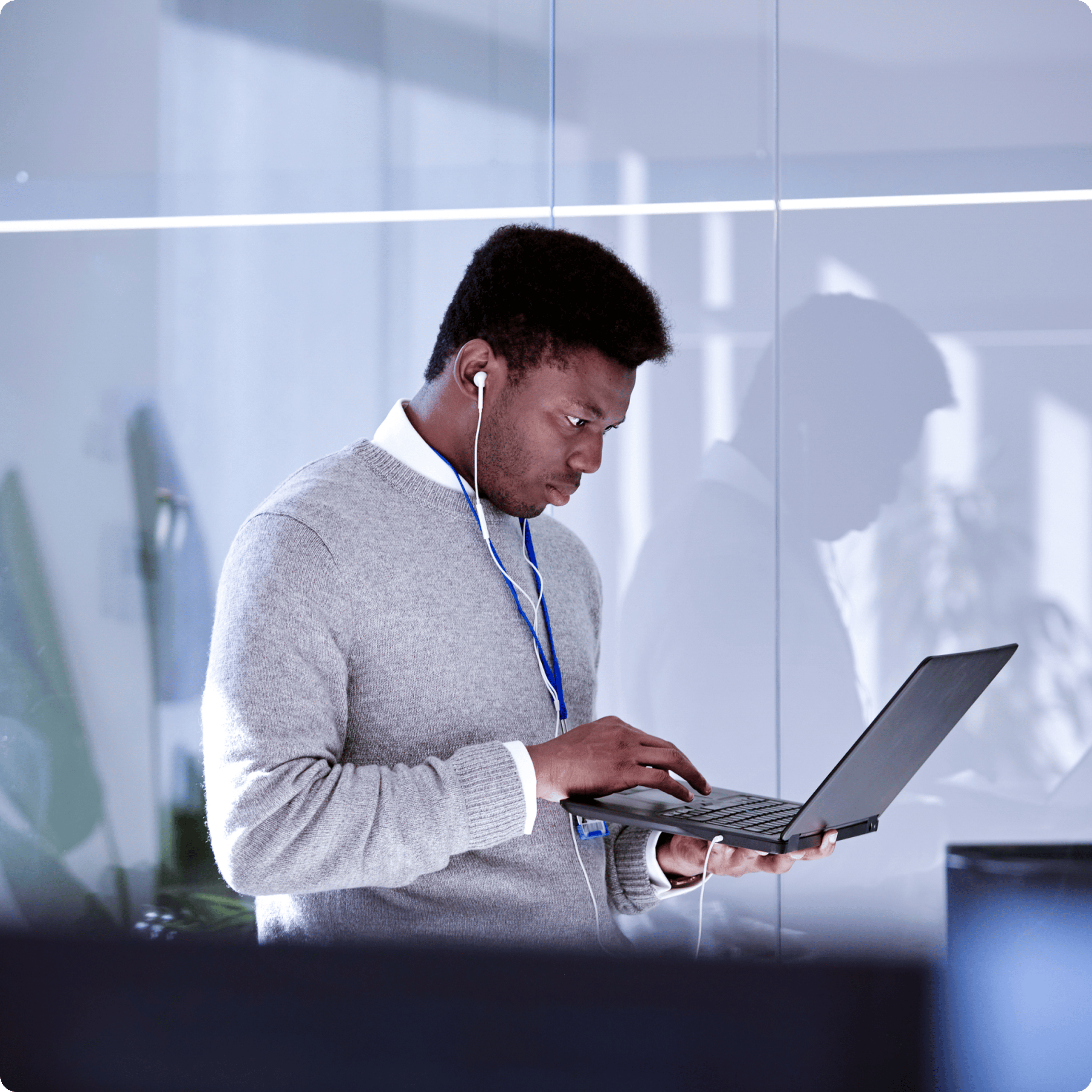 Man in a light office setting holding an open laptop and looking down at the screen, wearing wired earphones
