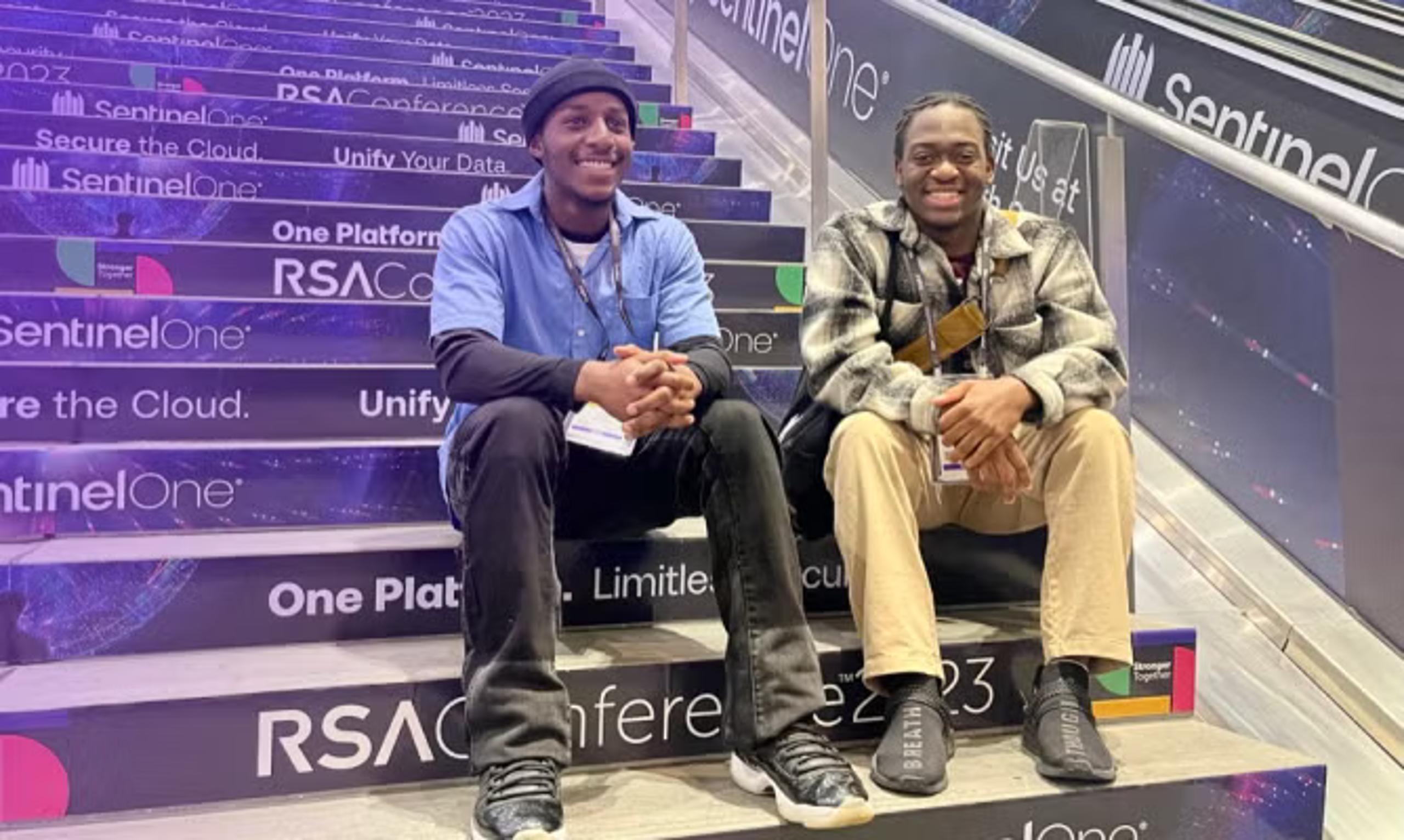 Two smiling people sit on purple branded staircase steps with visible text like SentinelOne, One Platform, RSA, Secure the Cloud, Unify