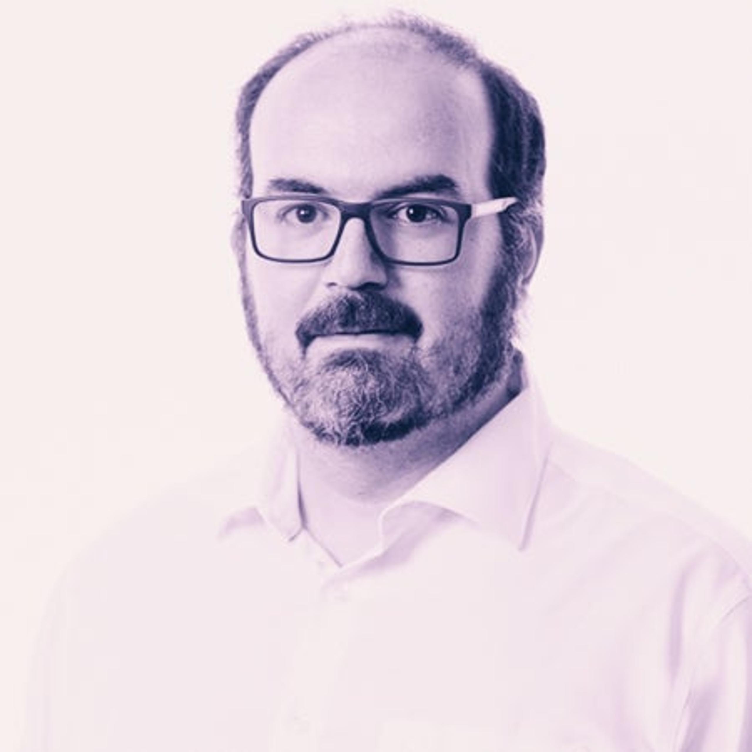 Chest-up portrait of an adult man with glasses, mustache and full beard, wearing a light collared shirt against a light background