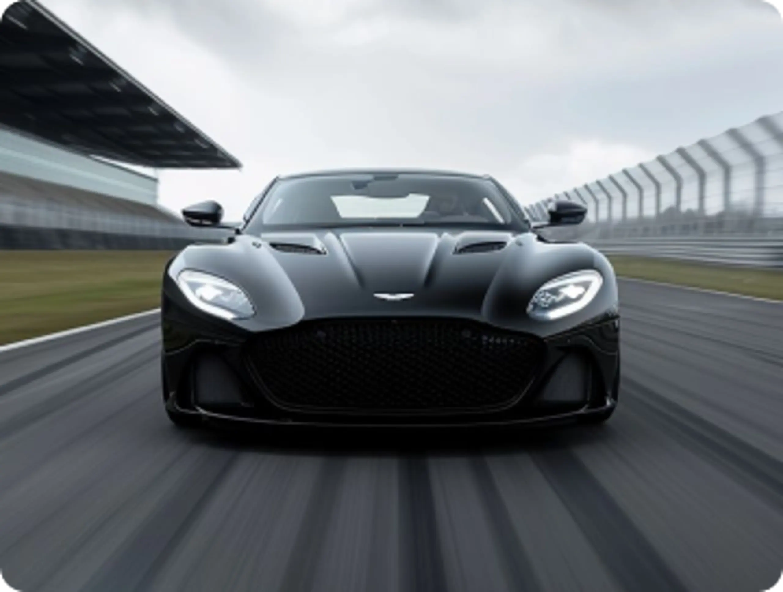 Front view of a glossy black sports car driving fast on a racetrack, with grandstand left, fence right, gray overcast sky