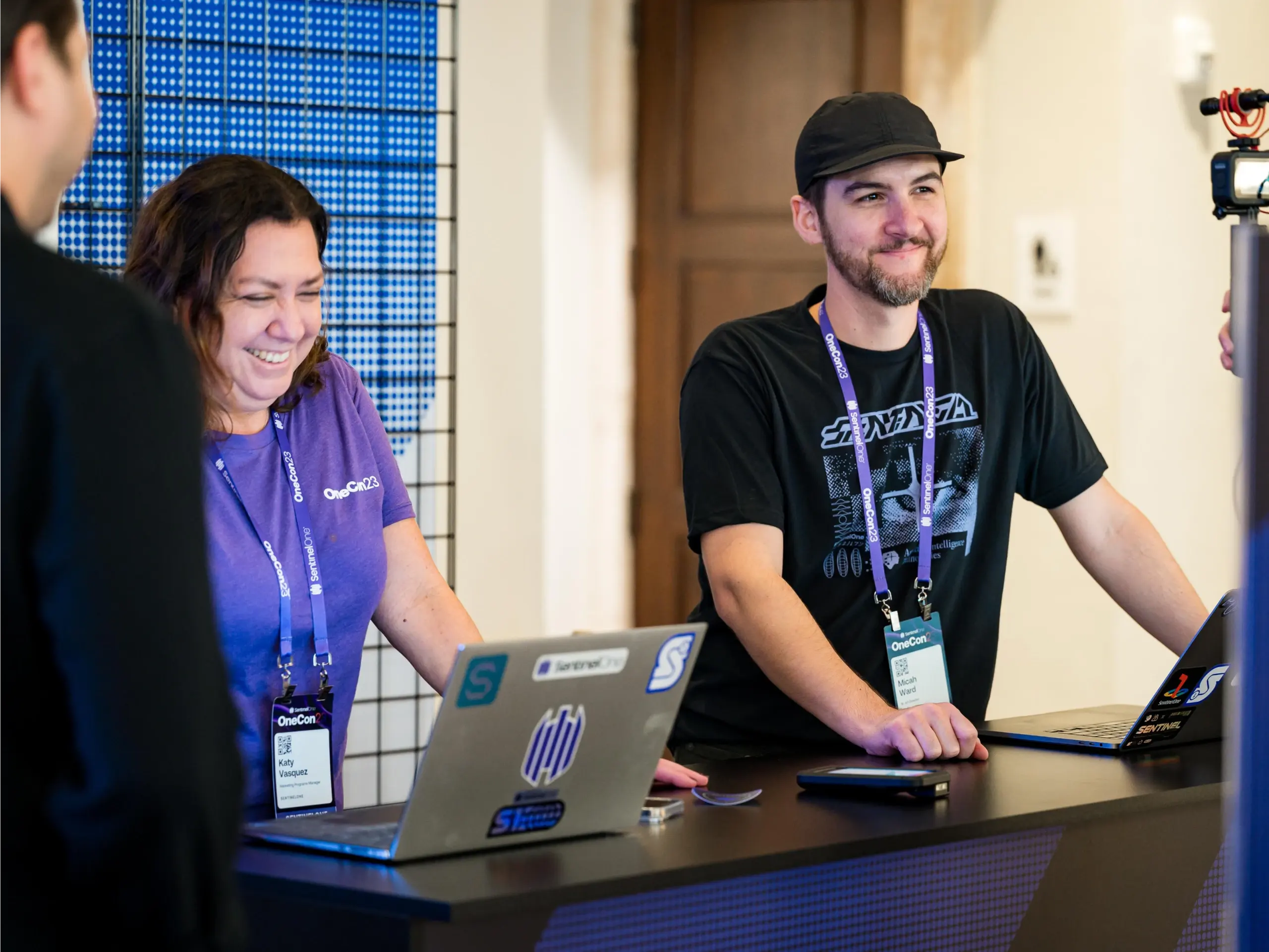 Two people with lanyard ID badges work at a table with open laptops, a smartphone nearby, and a blue grid panel in the background