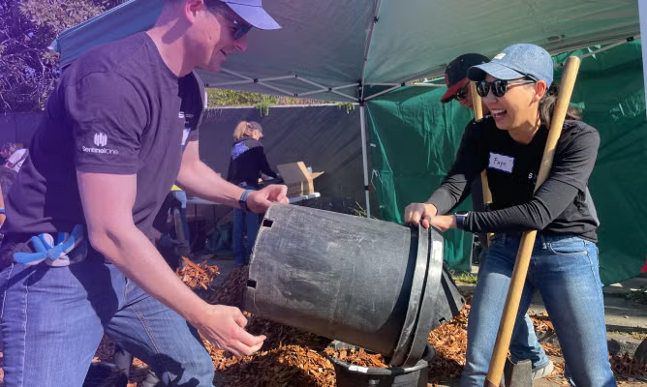 Two adults under a white canopy tent working together beside a tilted dark barrel; mulch on the ground and a name tag label on the woman