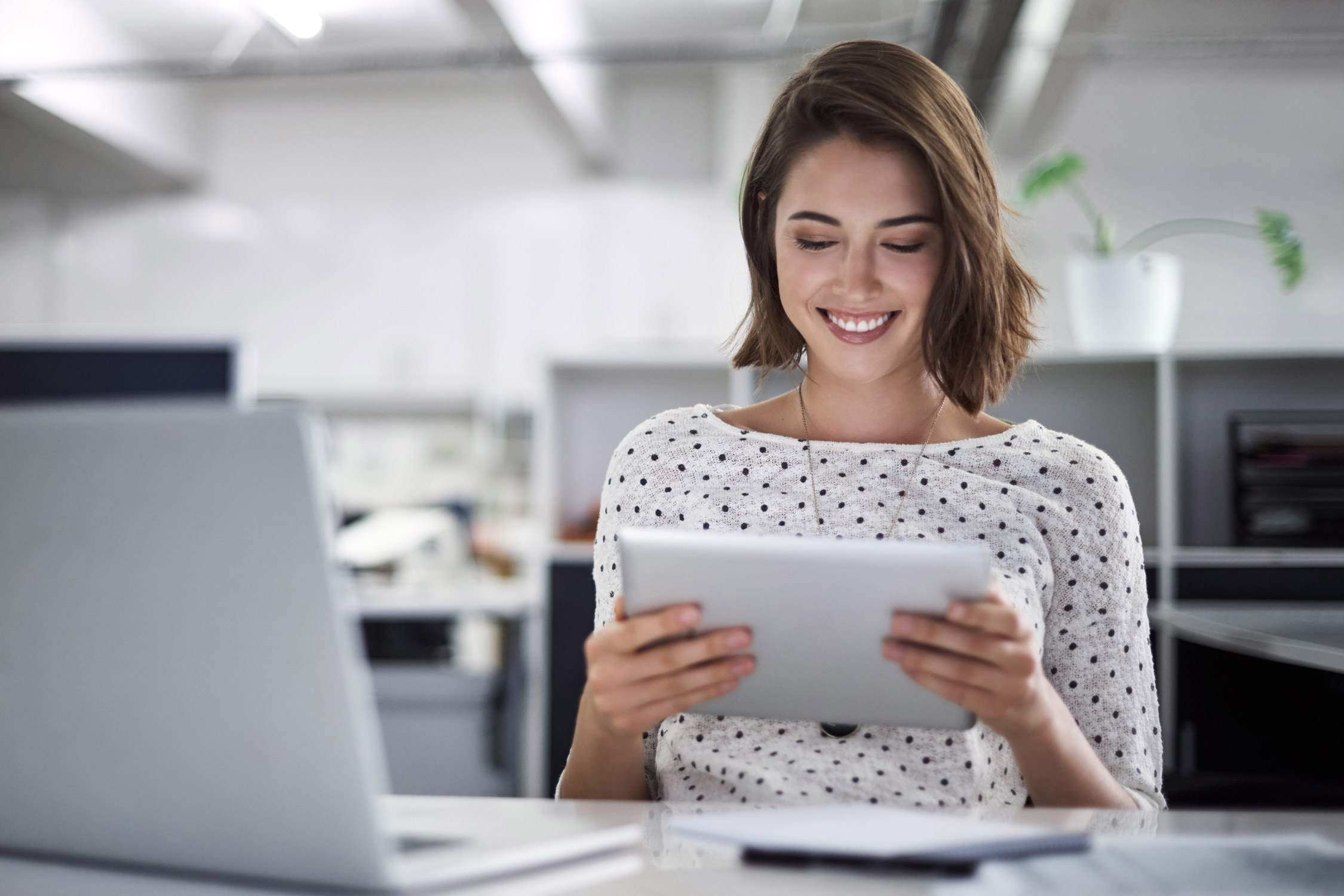 Frau mit gepunkteter Bluse lächelt an einem Schreibtisch mit einem Laptop und Tablet