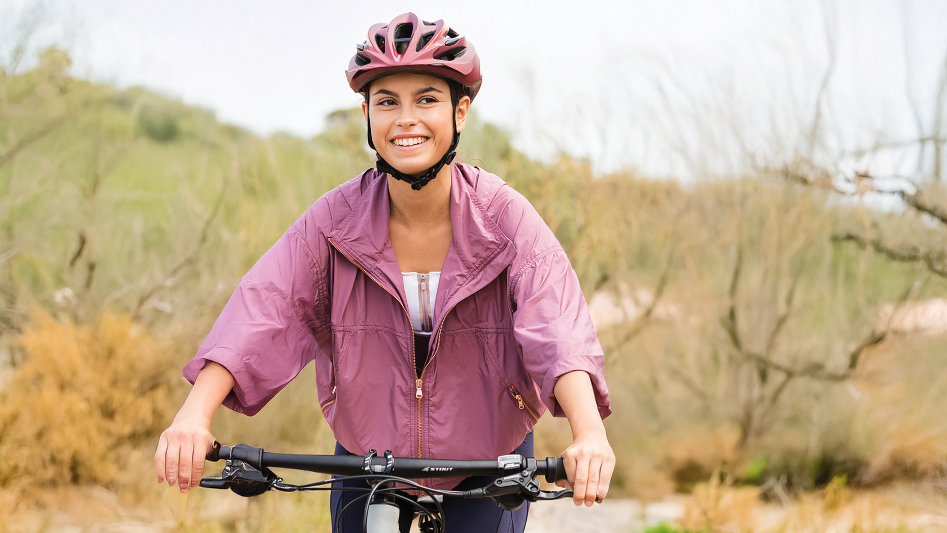 امرأة ترتدي ملابس رياضية وردية، تبتسم، تركب دراجة مع خوذة للسلامة