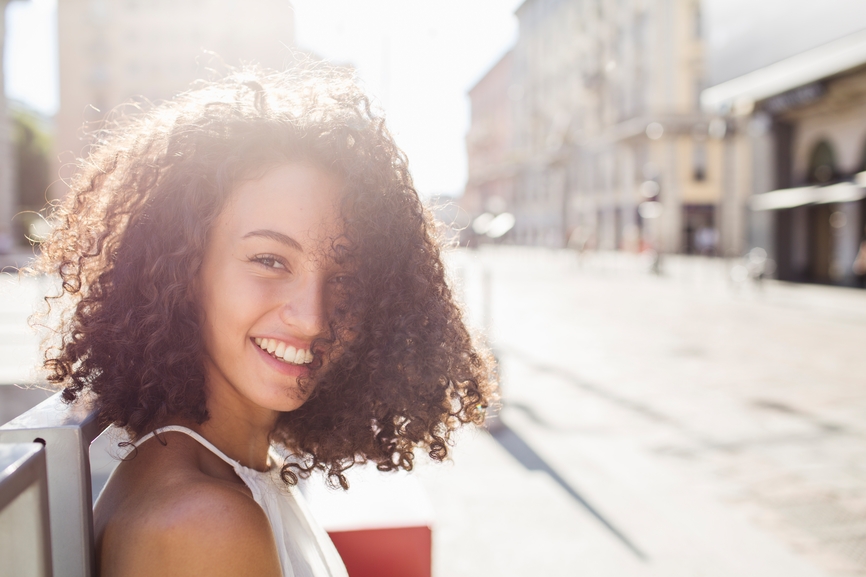 A young curly haired girl looking over her shoulder and smiling with the sun in the background.