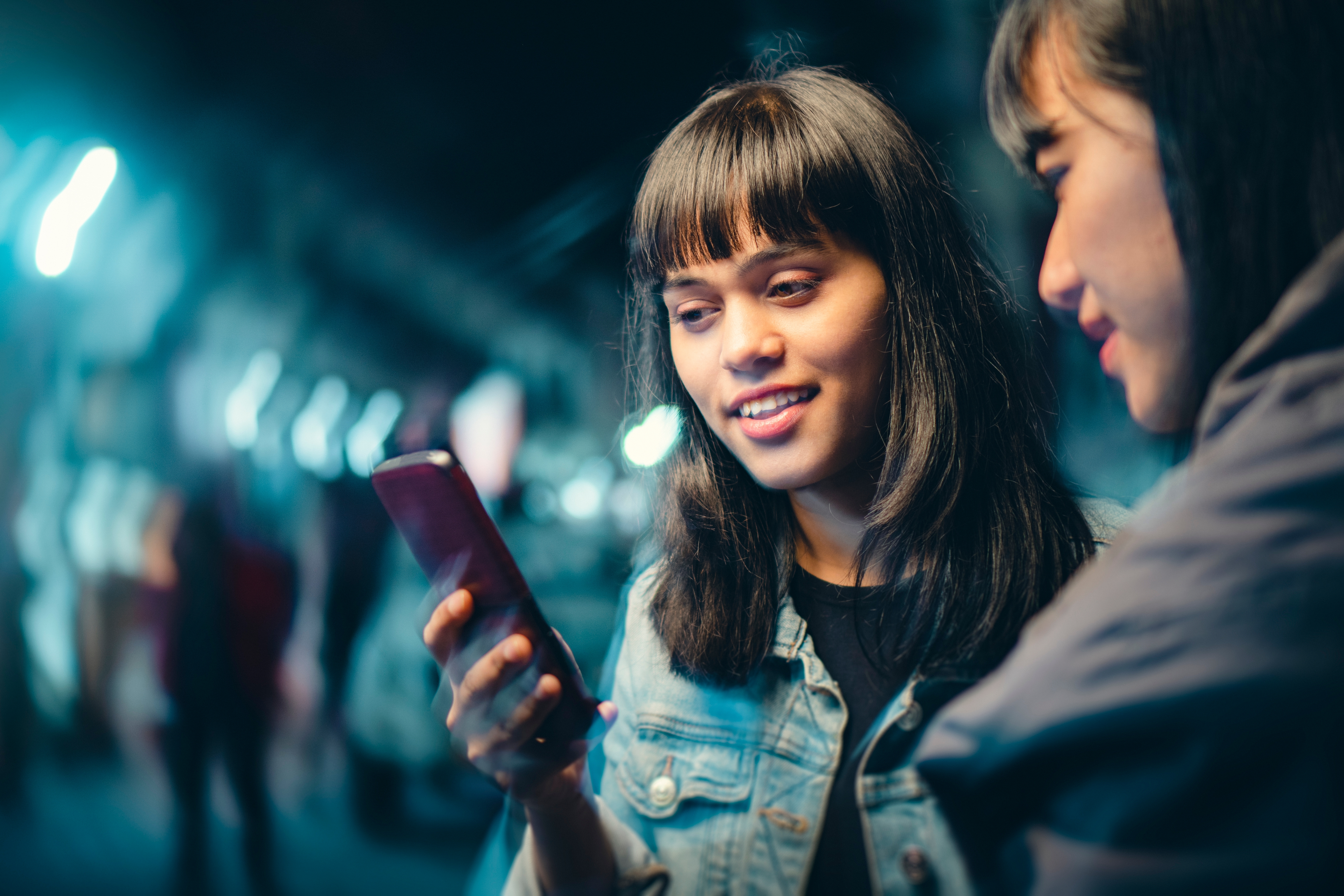 Outdoor image of happy Asian, Indian multiethnic female friends gossip and use smartphone together on city street at night.
