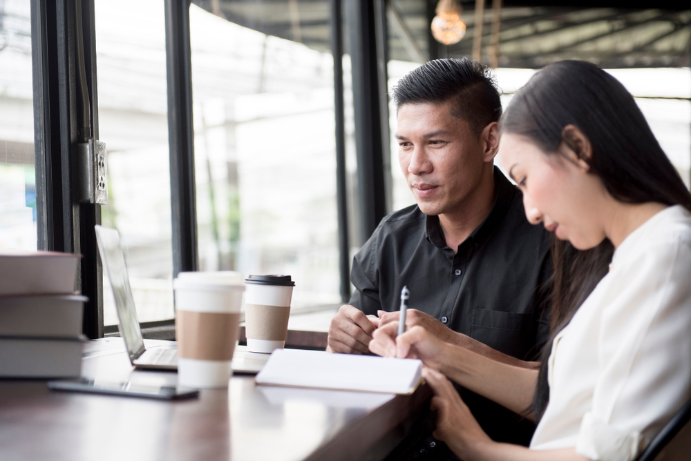 Smiling young woman writing notes and a focus young man looking at a laptop