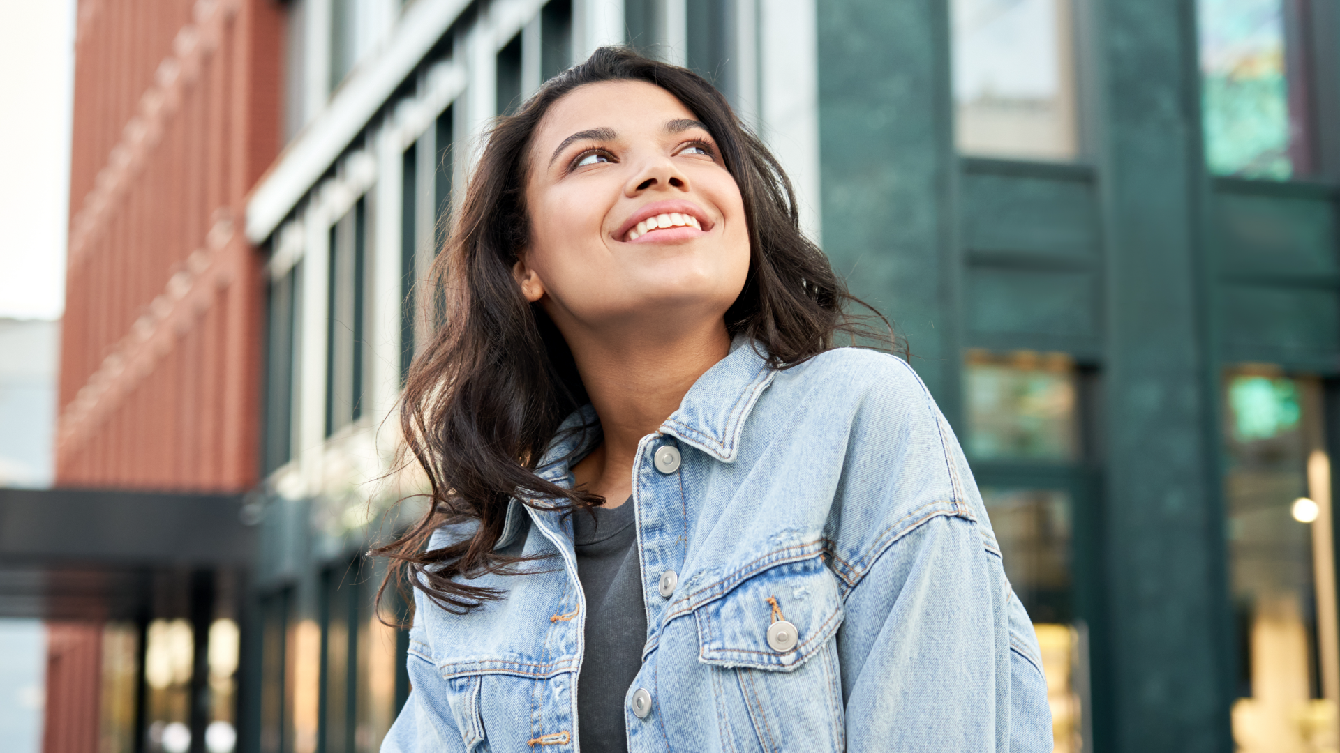 امرأة شابة سمراء ترتدي قميص جينز تقف في زاوية بالخارج، تبتسم وتنظر للأعلى بتعبير مليء بالأمل