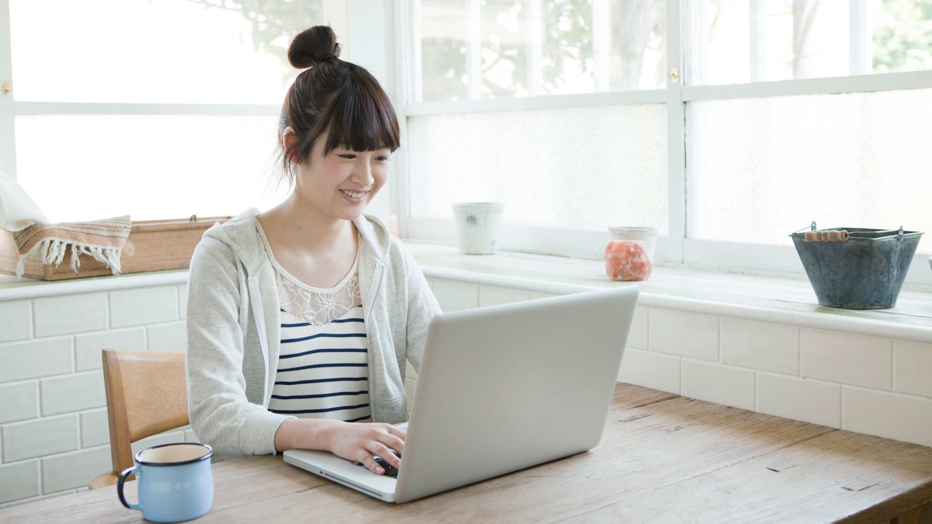 Young Asian woman on her laptop wearing a gray cardigan over a striped shirt.