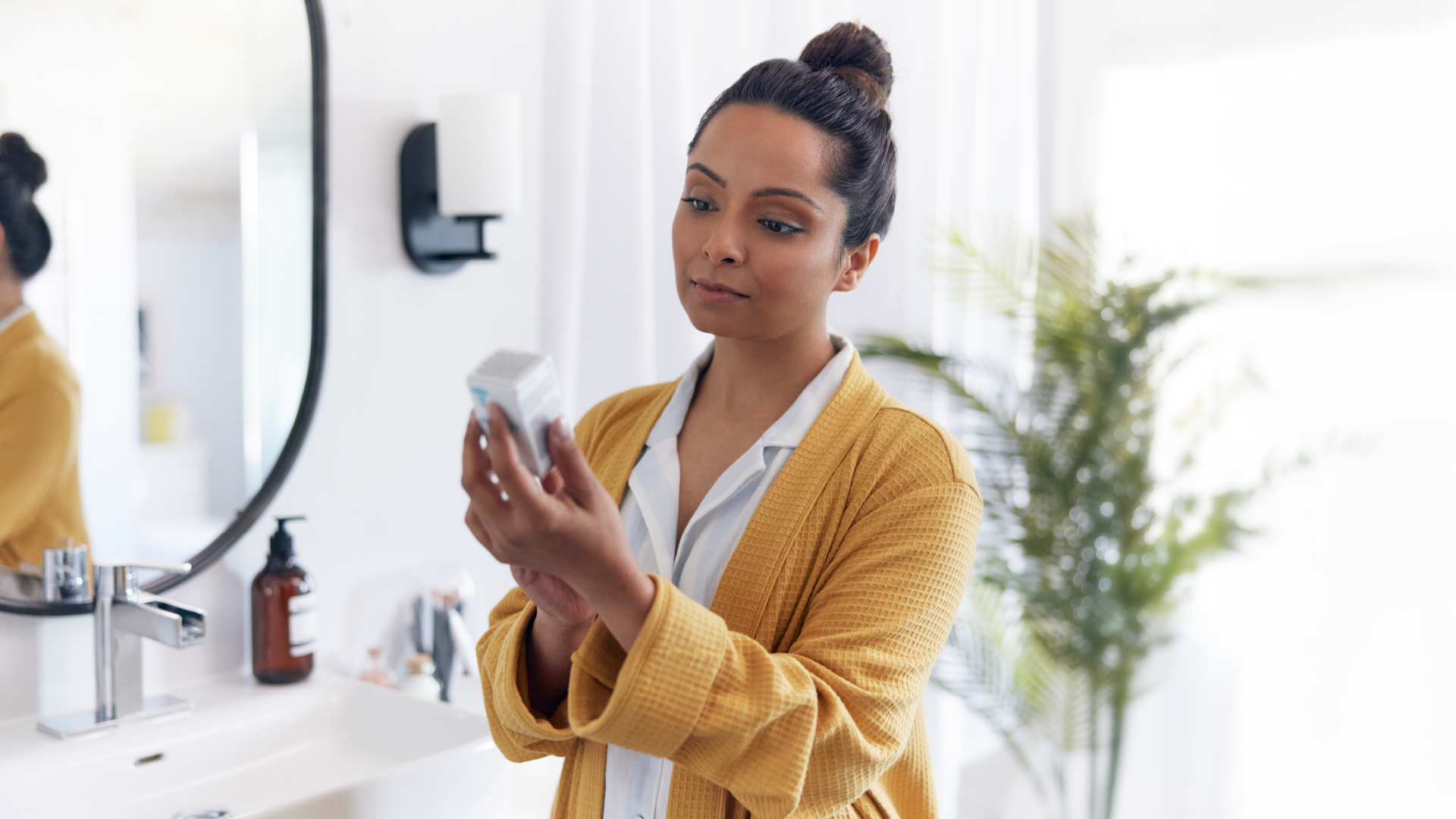 امرأة من أصل عرقي ترتدي بدلة صباحية برتقالية تحمل صندوقًا وتحاول قراءة نص صغير أمام مرآة الحمام