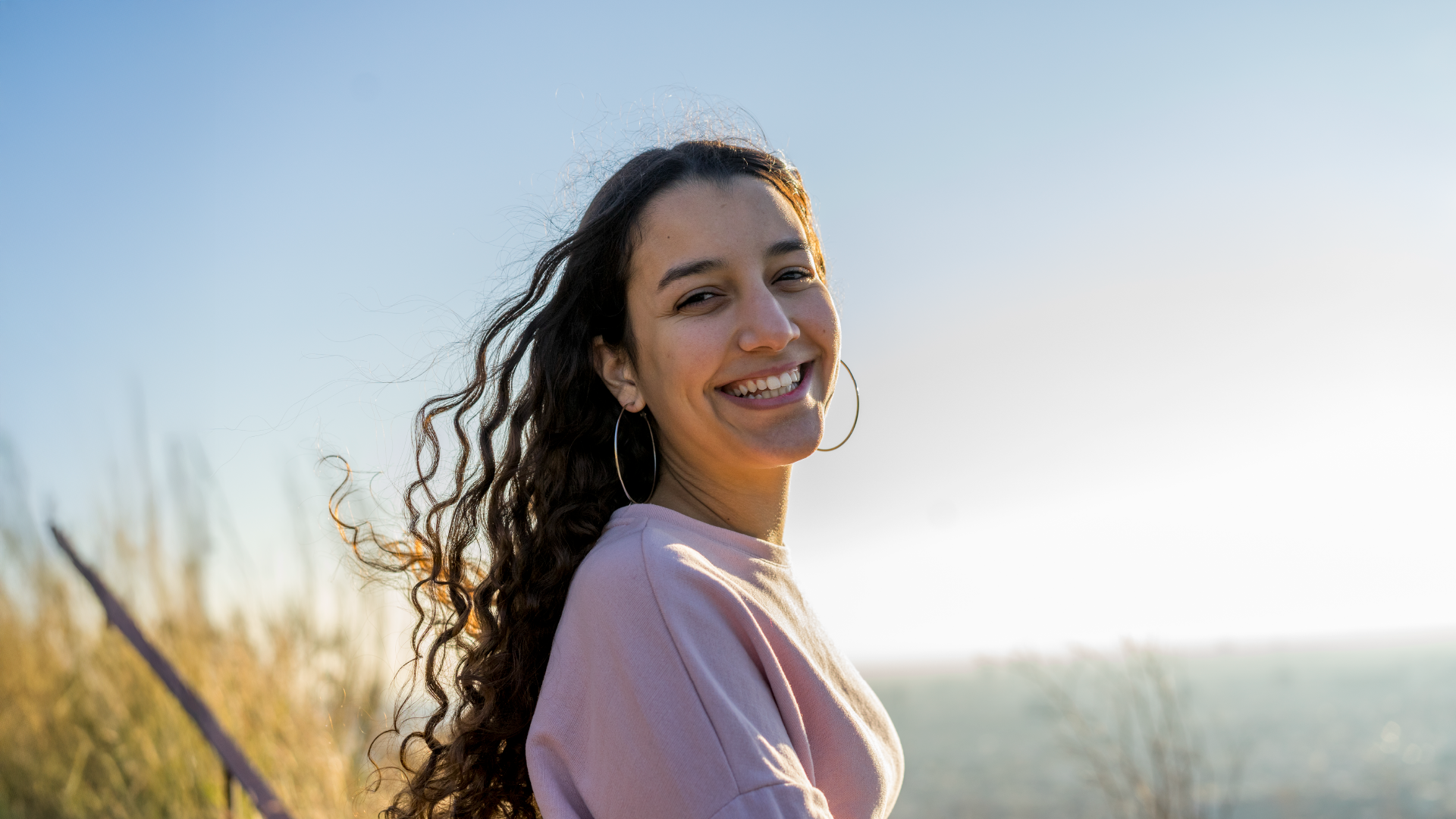 امرأة شابة ذات شعر مجعد تبتسم للكاميرا فوق كتفها بجانب البحر، مع أشعة الشمس تشرق من اليمين