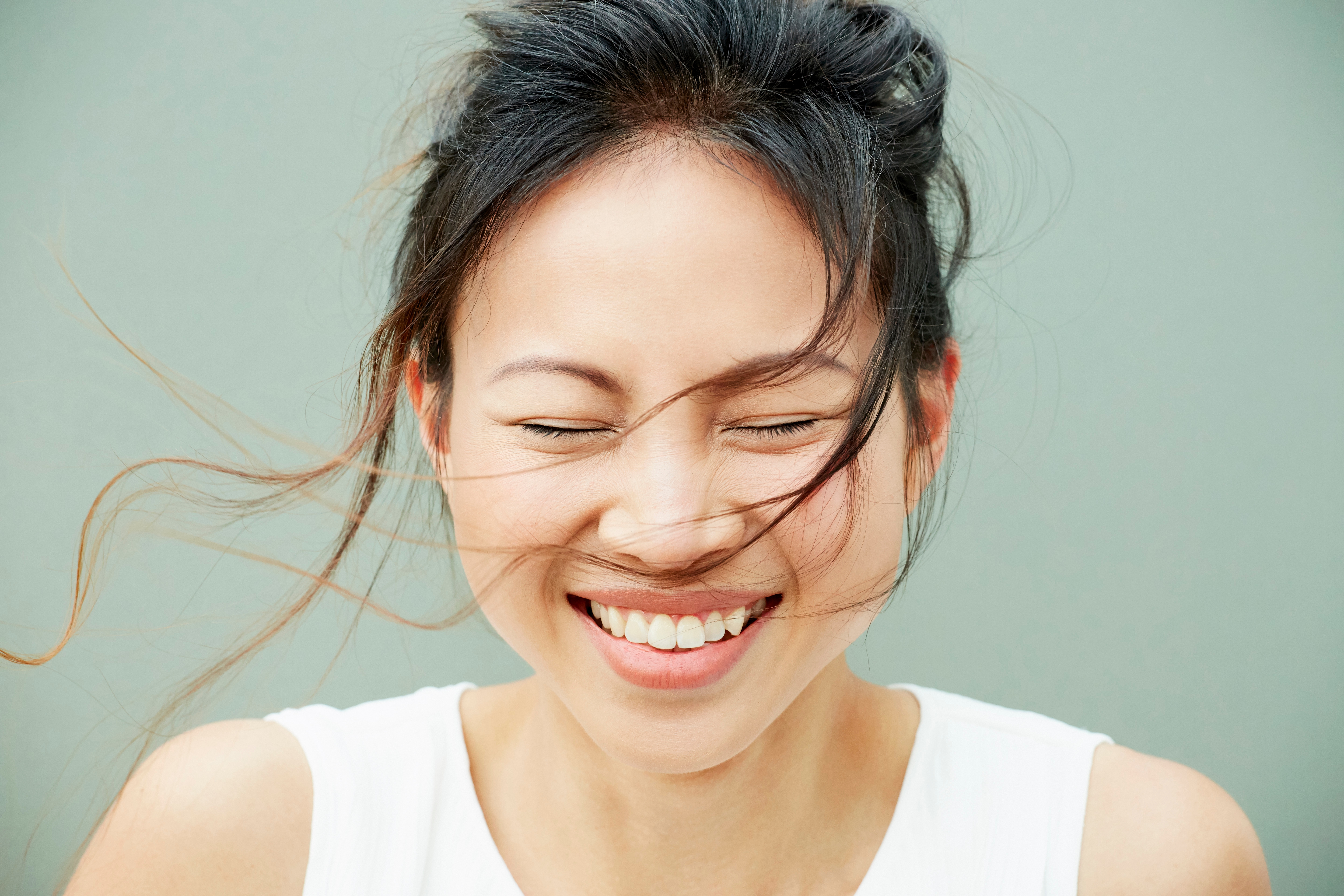 A woman in white tank top closing her eyes with a wide smile on her face looking at the front