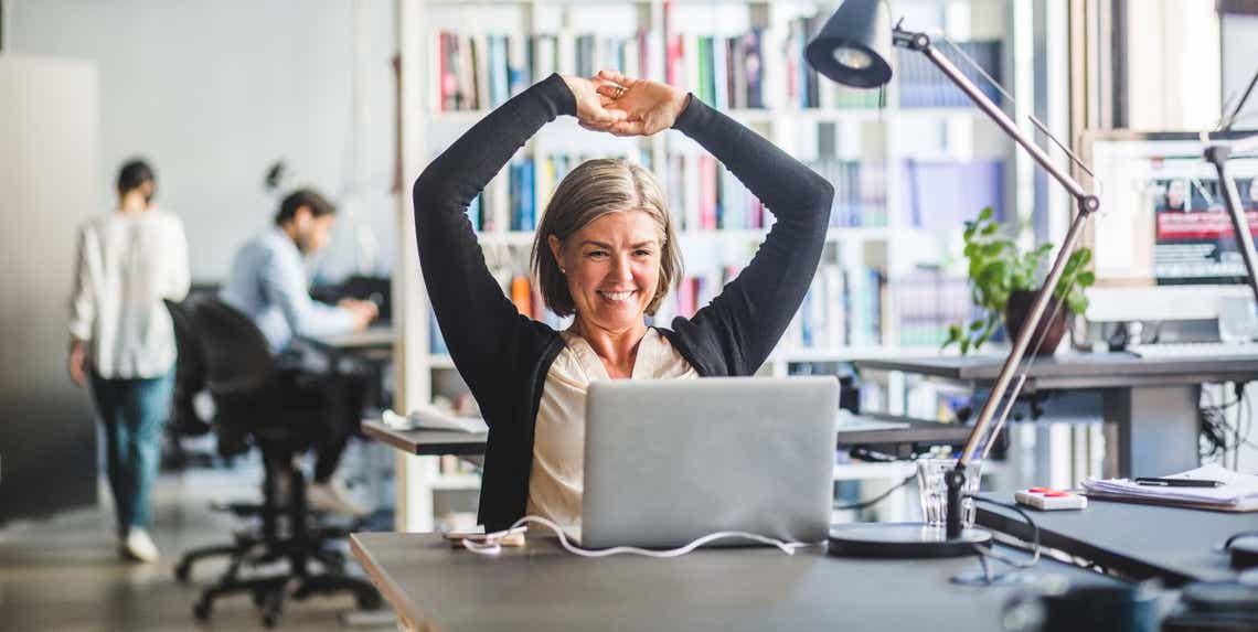 sales enablement professional sitting at her computer in a busy office with her hands in the air celebrating the performance of the sales team she supports.
