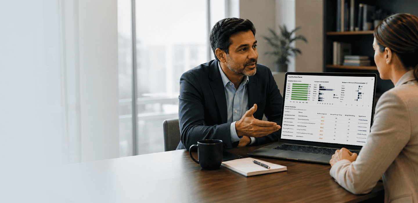 A sales manager talking with a seller across a desk, an Accelerate Prism capability dashboard open on the laptop between them