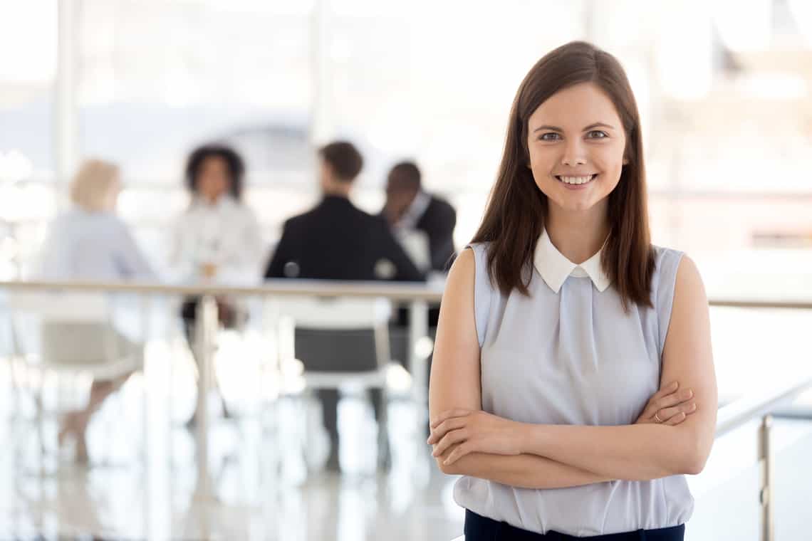 female sales manager looking confidently into the camera while her team works in the background behind her. The team is working hard because she has developed the sales manager traits that build high performing teams.