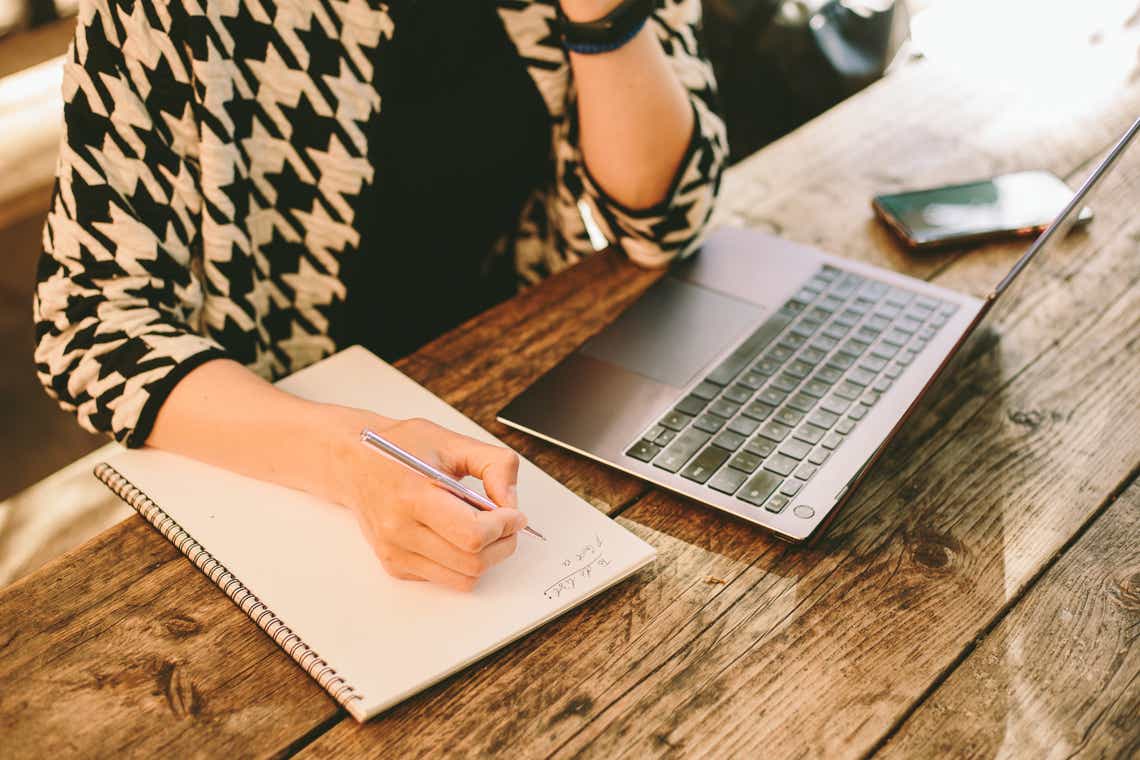 A person creating a to-do list in notebook next to computer for sales accountability