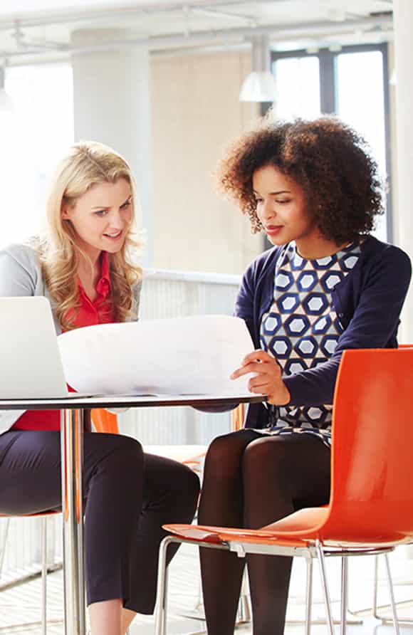 two professional women seated at a table in a sunny office building with an open laptop on the table looking as a document.