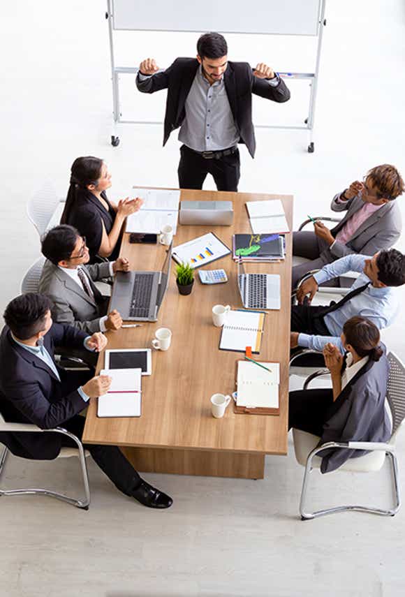 group of sales people sitting at a table looking at a free-standing white board engaged in a meeting lead by a confident leader.