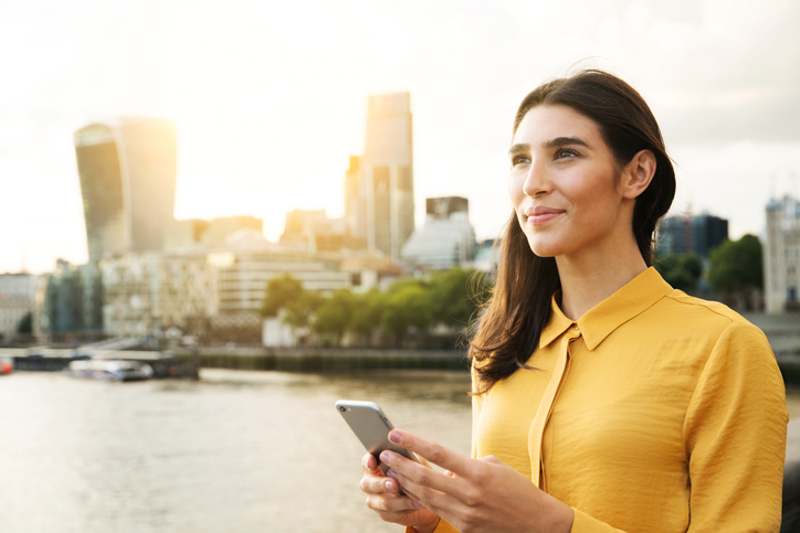 young business woman standing with a phone with a river and city line in the background looking ahead to the future.