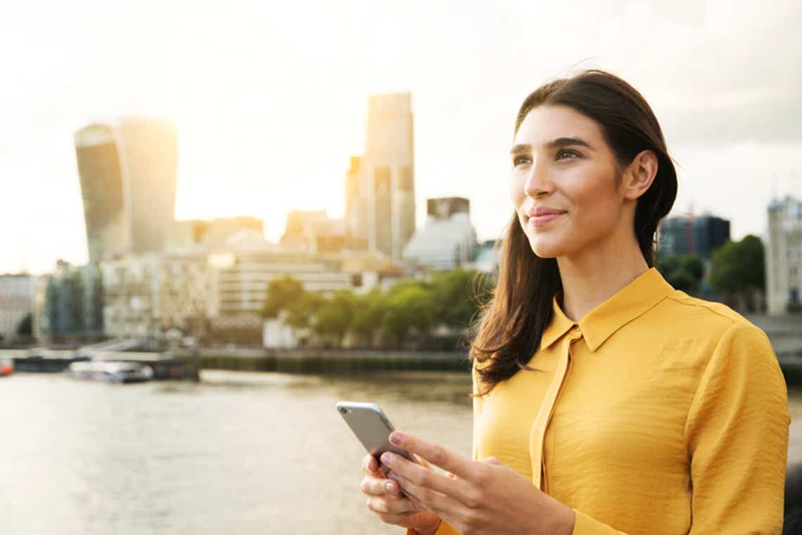 young business woman standing with a phone with a river and city line in the background looking ahead to the future.
