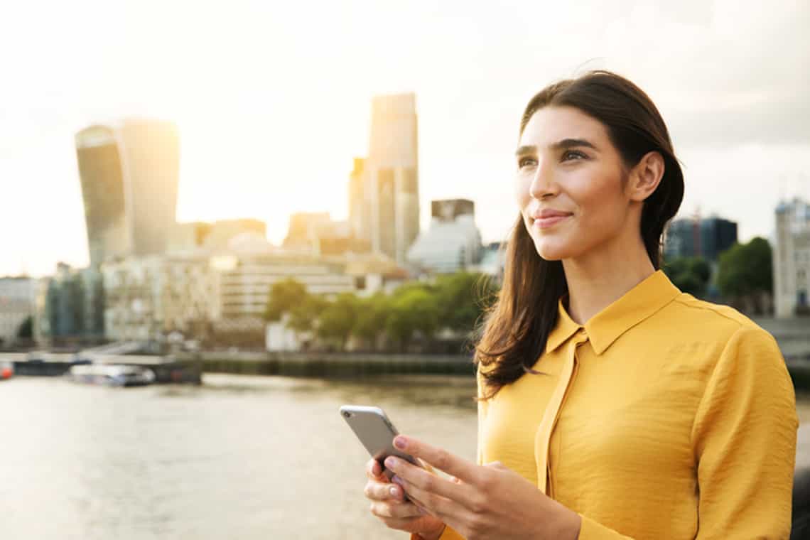 young business woman standing with a phone with a river and city line in the background looking ahead to the future.