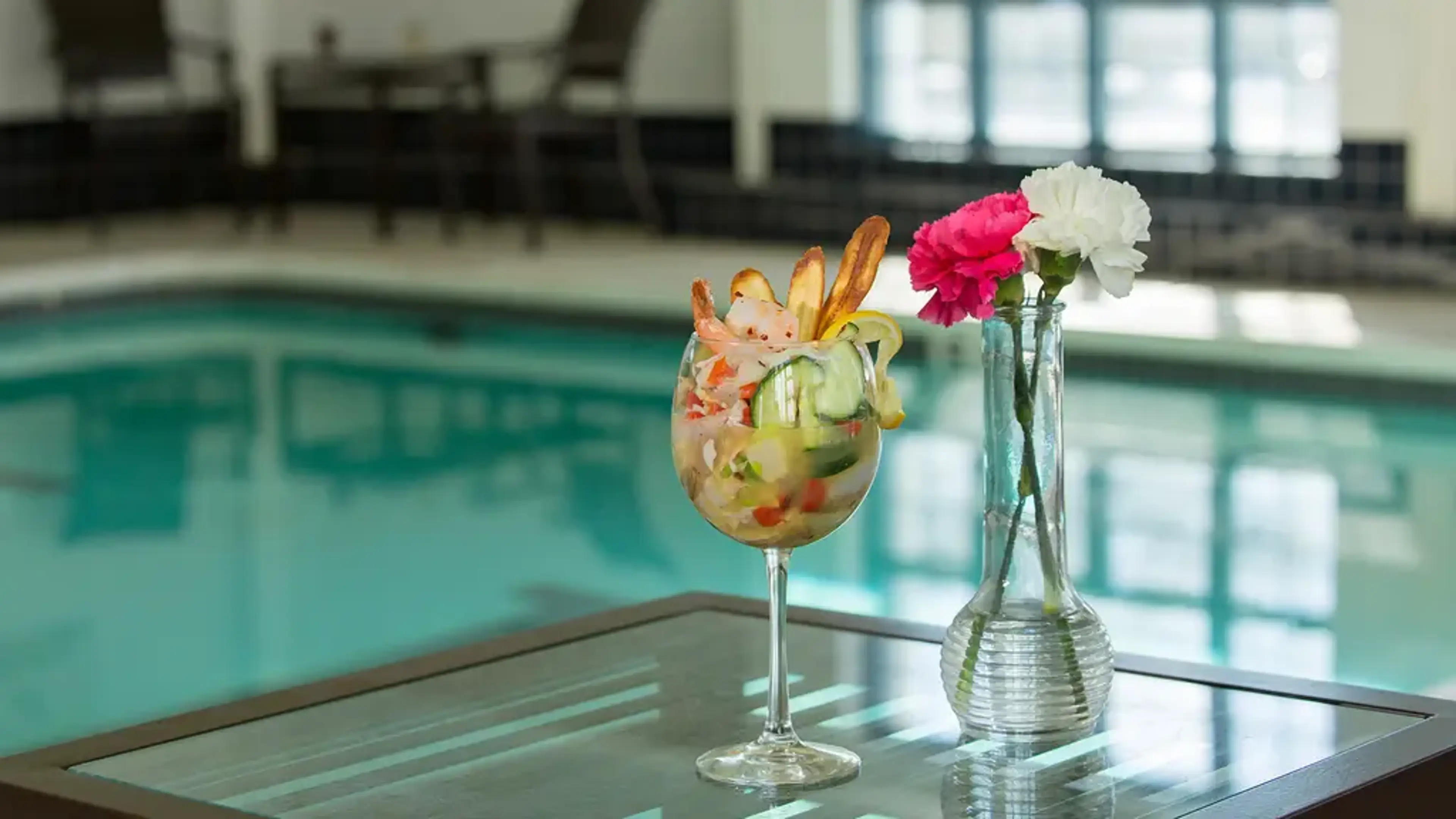 A full wine glass with fruits and flowers with a red flower in a vase next to it.