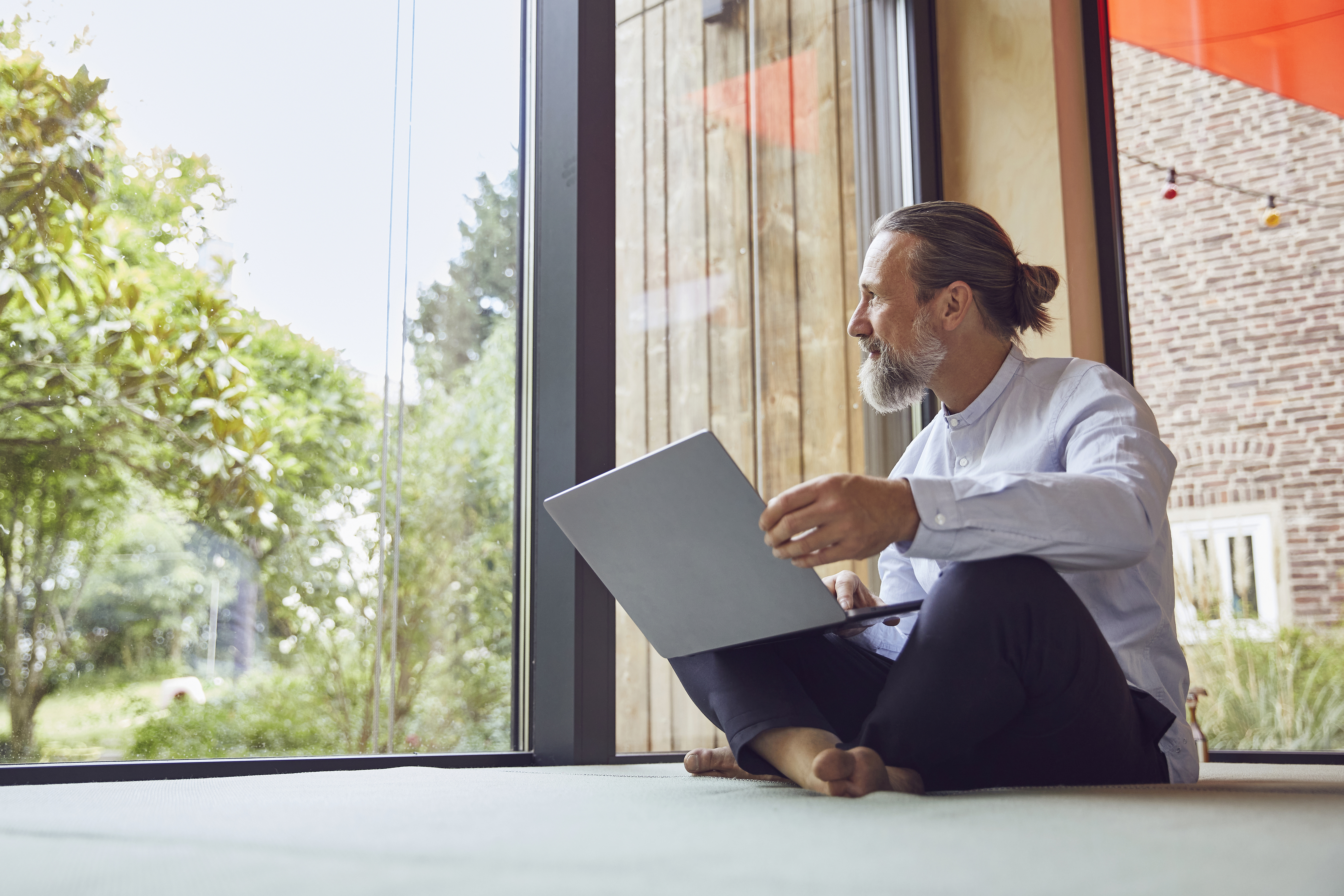 Ein Mann sitzt mit dem Laptop auf dem Schoß vor einem großen Fenster.