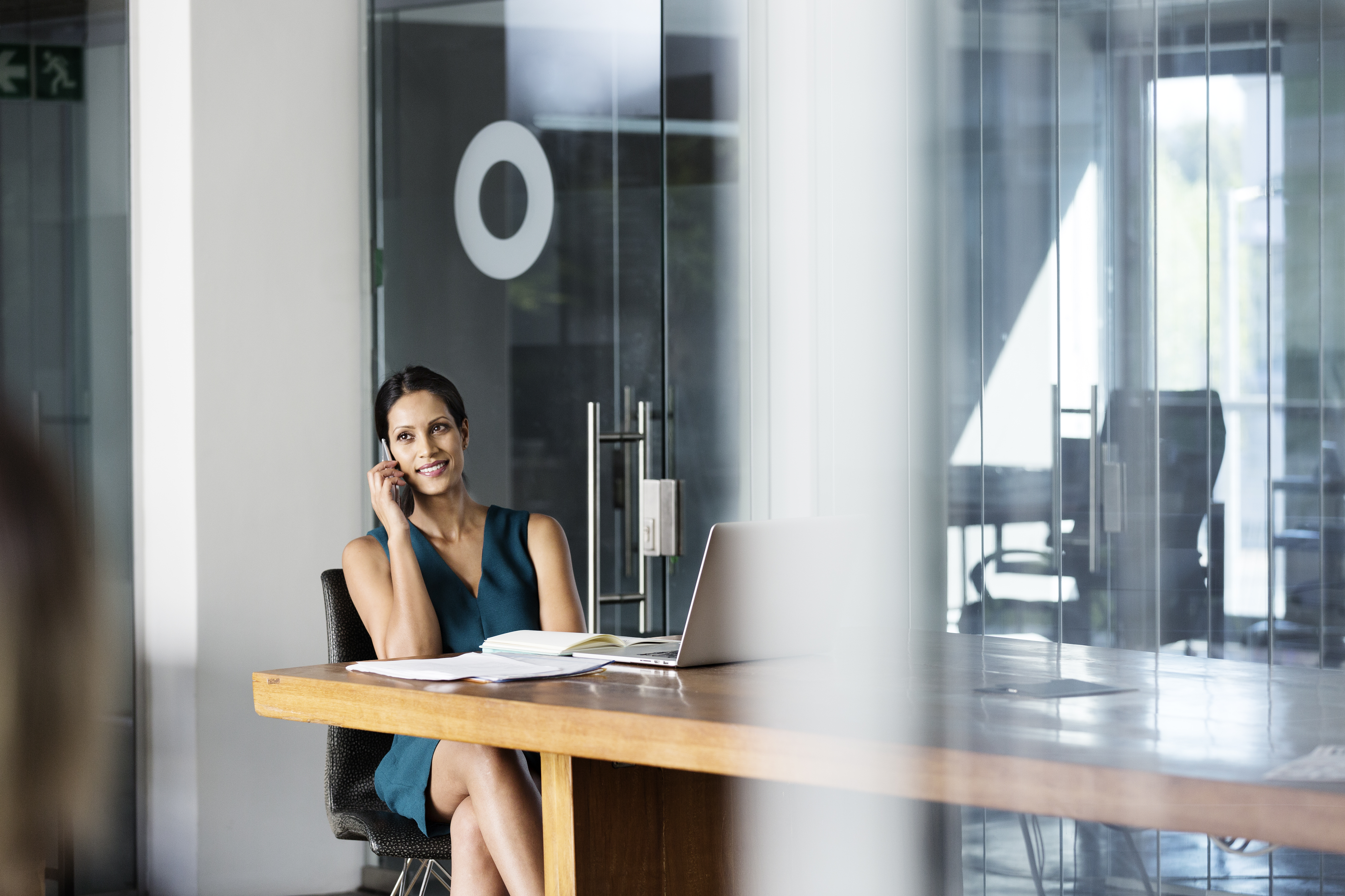 Eine Frau sitzt am Tisch mit Laptop und Arbeitsbüchern und telefoniert.
