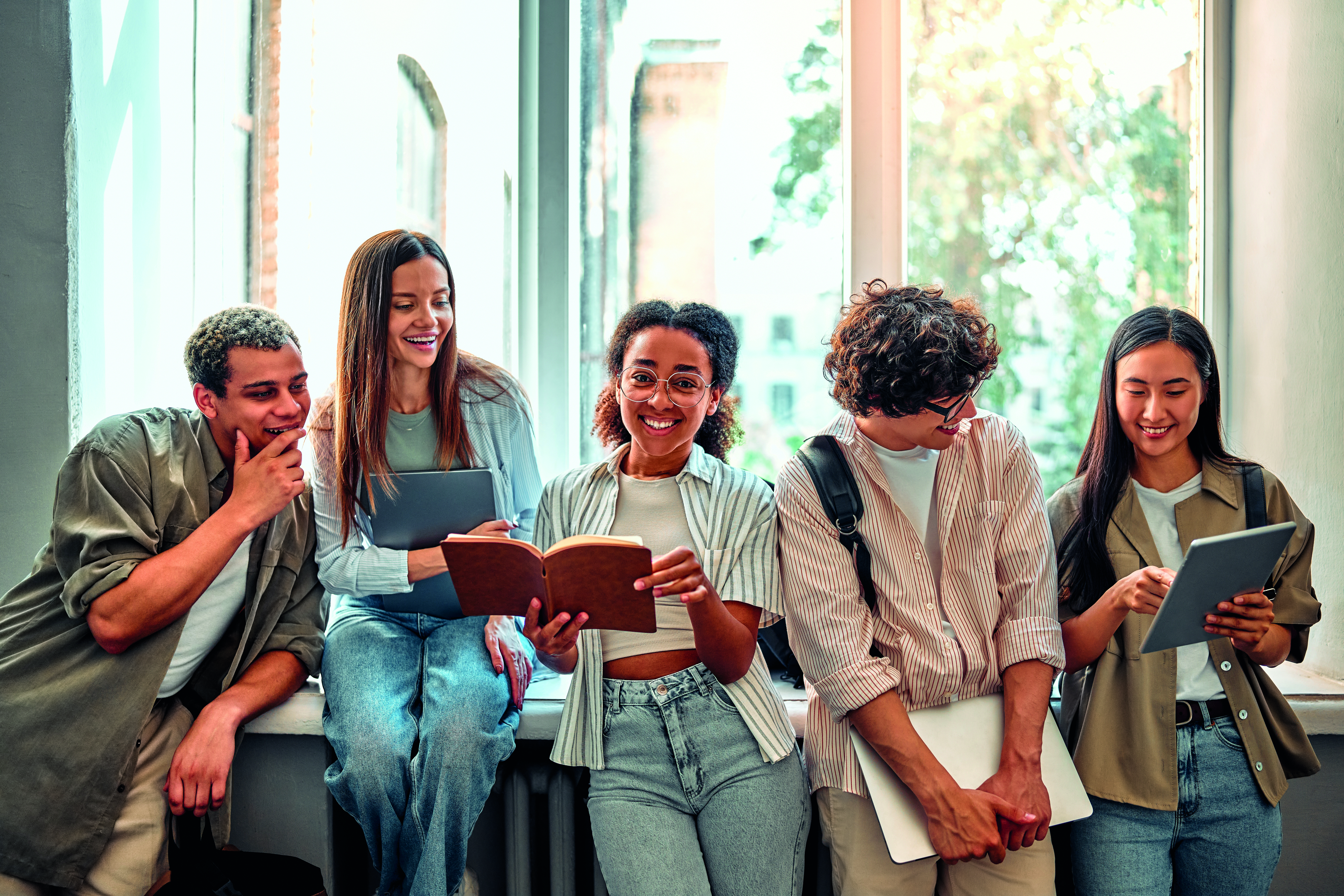 Gruppe von Jugendlichen stehen oder sitzen vor einem Fenster mit Büchern in der Hand und lächeln