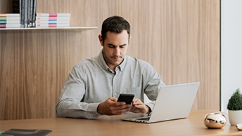 Wide|Business man in an office working on a laptop and viewing his smartphone