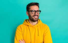 A smiling man in a yellow hoodie with his arms crossed against a turquoise background.